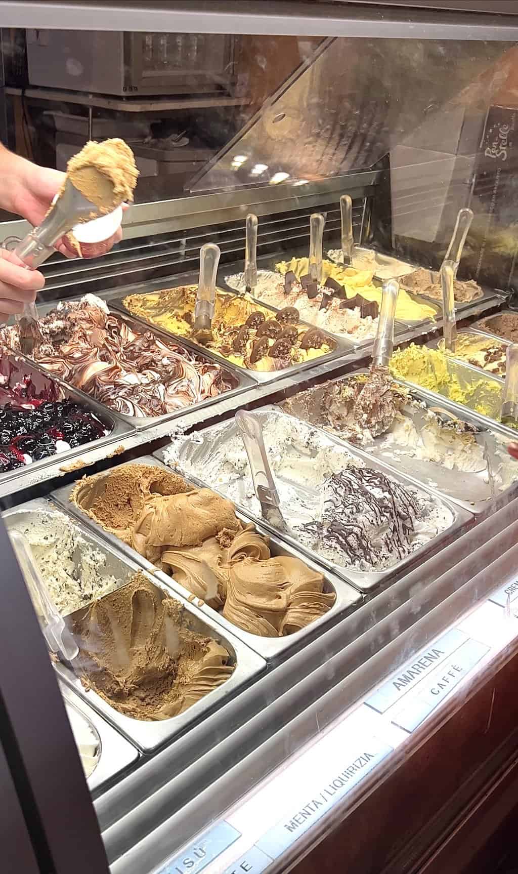 A photo of a selection of Gelato available at Frigidarium, one of the best Gelato venues in Rome. A hand extends out in the top left corner to scoop a coffee flavoured gelato into a cup. 