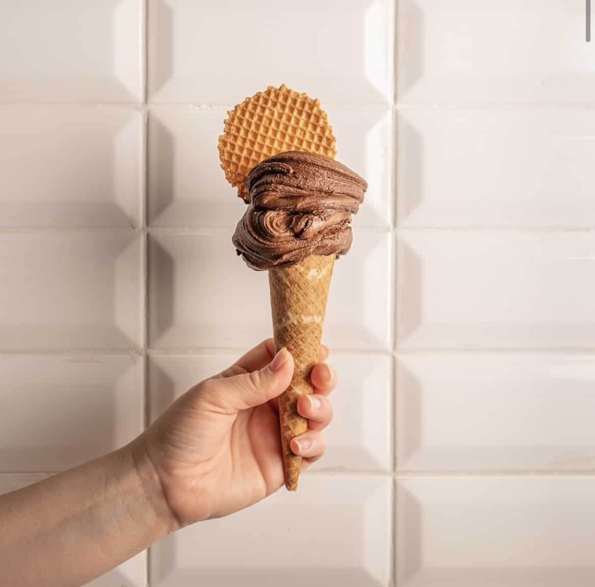 A side on image of a waffle cone with chocolate ice cream held against a white tiled wall at Come Il Latte in Rome. The ice cream is topped with a extra waffle. 