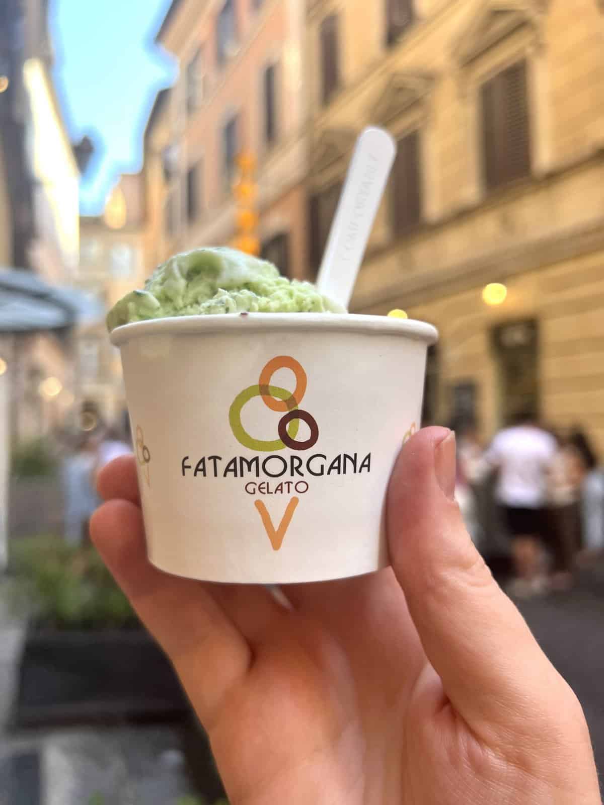 A hand holds out a white cup filled with basil and walnut gelato from Frigidarium, a gelato shop in Rome. The background is a Roman streetscape during the afternoon. 