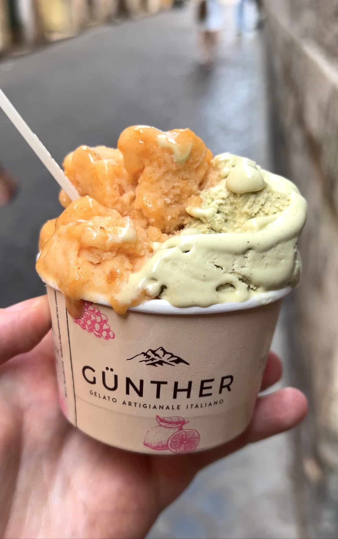 An image of a hand holding a cup of gelato from Günther gelato in Rome. The cobbled streets of Rome are in the background, and the melty pistachio and apricot gelato is in the foreground.