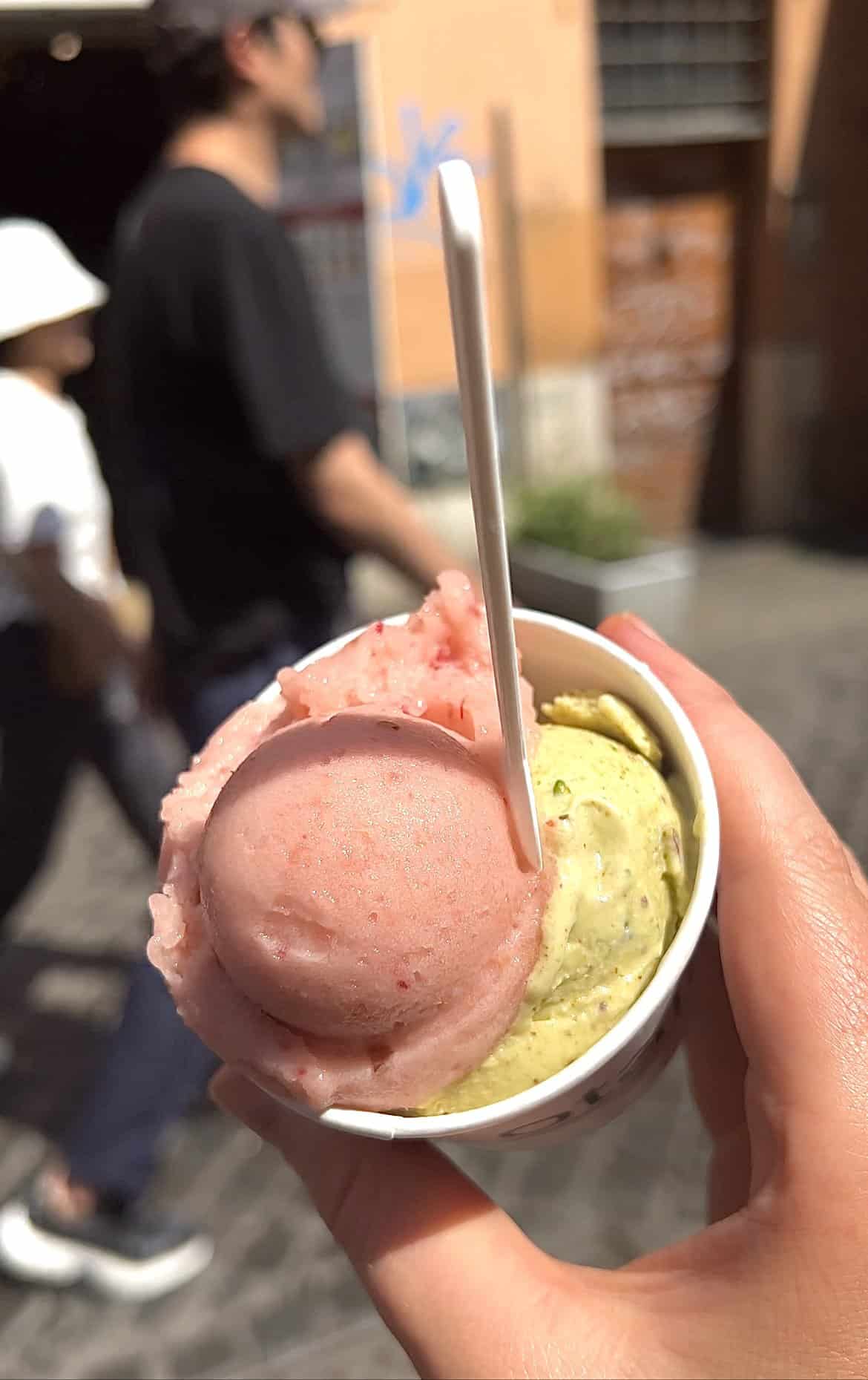 An image of an ice cream cup filled with peach and pistachio gelato from Otaleg in Trastevere, Rome. The scone is held by a female hand in the foreground, while a Roman streetscape and people walking are in the background. 