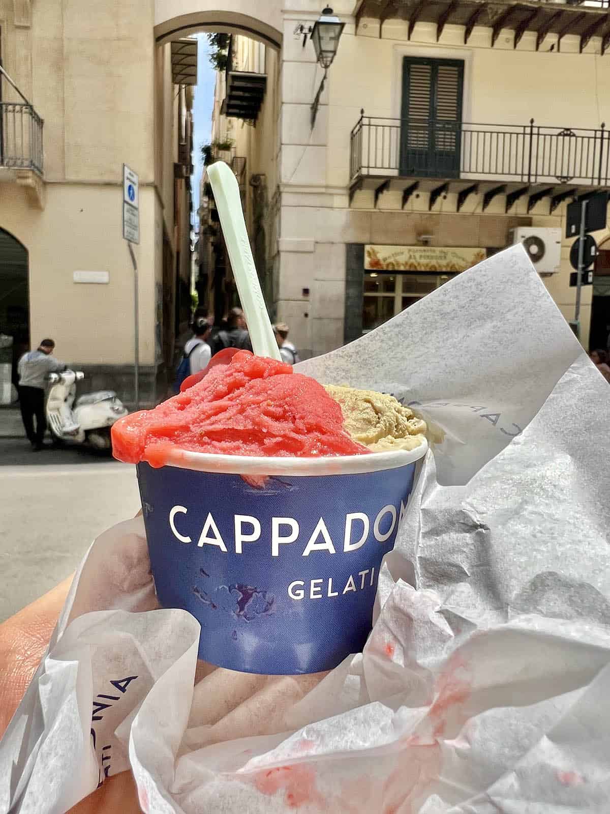 An image of a hand holding out a cup of gelato from Cappadonia Gelati in Palermo, Sicily. The gelato is strawberry and pistachio flavoured and it is in the foreground of a Palermo streetscape. 