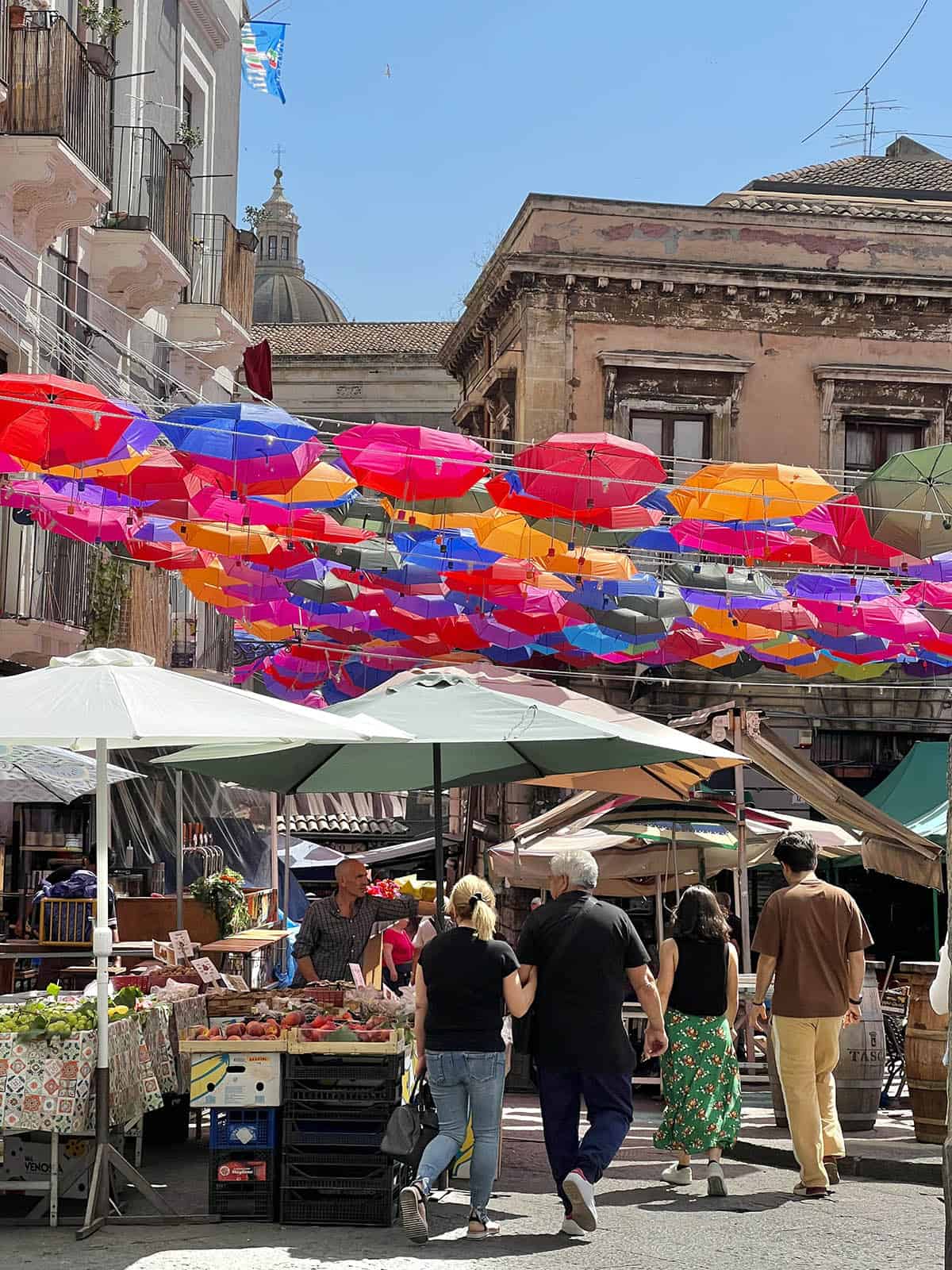 An image of the markets in Catania, Sicily. Bright coloured umbrella decoration shades the people and produce below, and the spire of the duomo sticks out in the background.