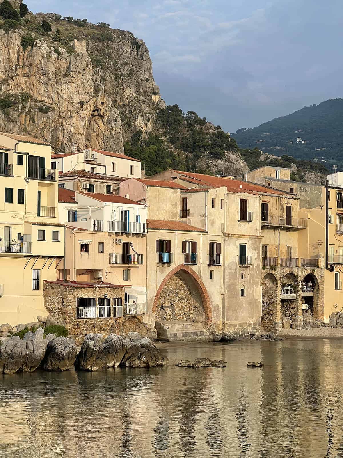 An image of the houses of Cefalu set against the rocky background. The ocean is the foreground, reflecting the orange hues of the buildings. 