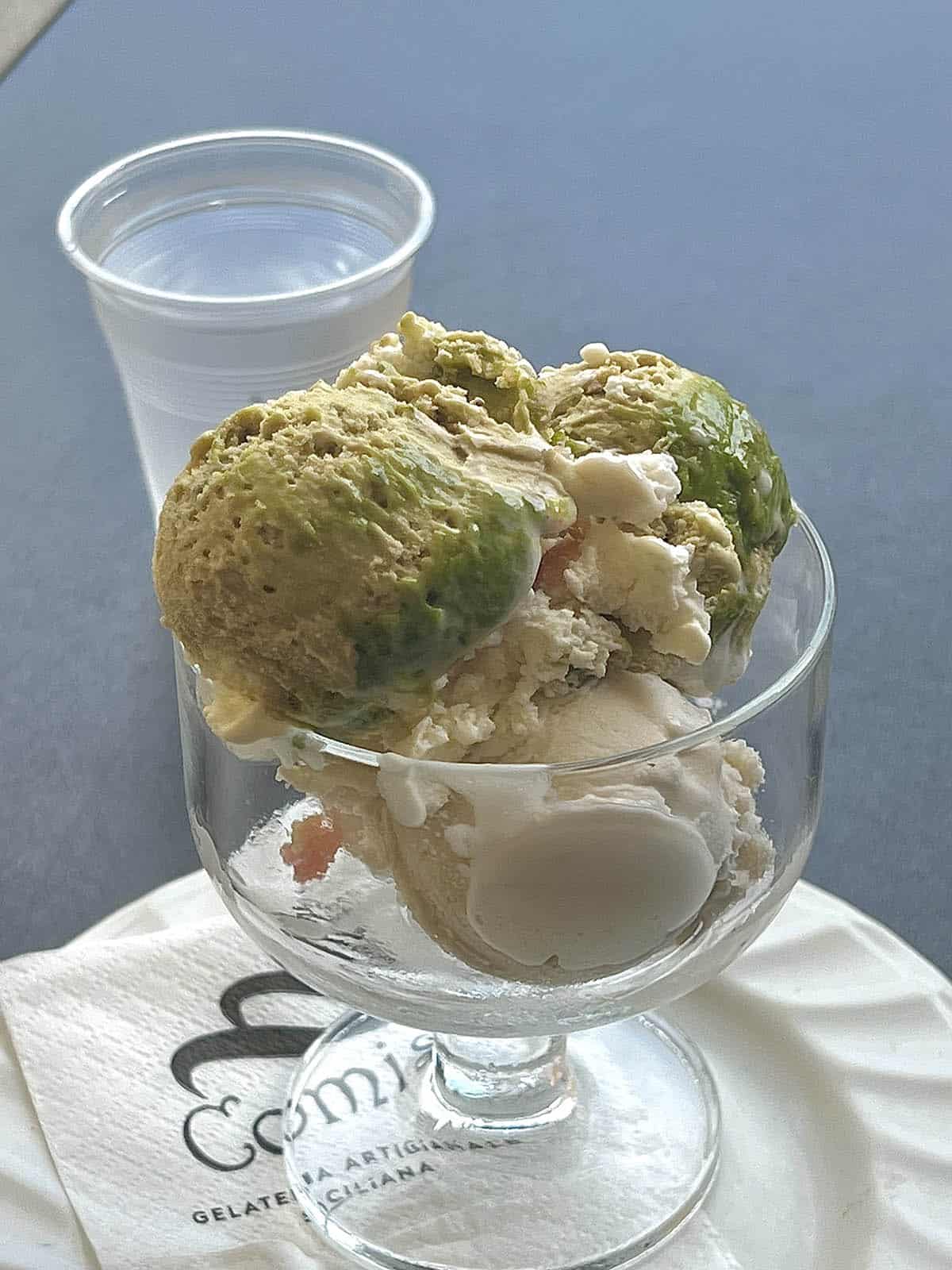 An image of a dessert glass filled with ice pistachio and lemon gelato. The glass sits on a branded serviette atop a plate on a dark grey table.