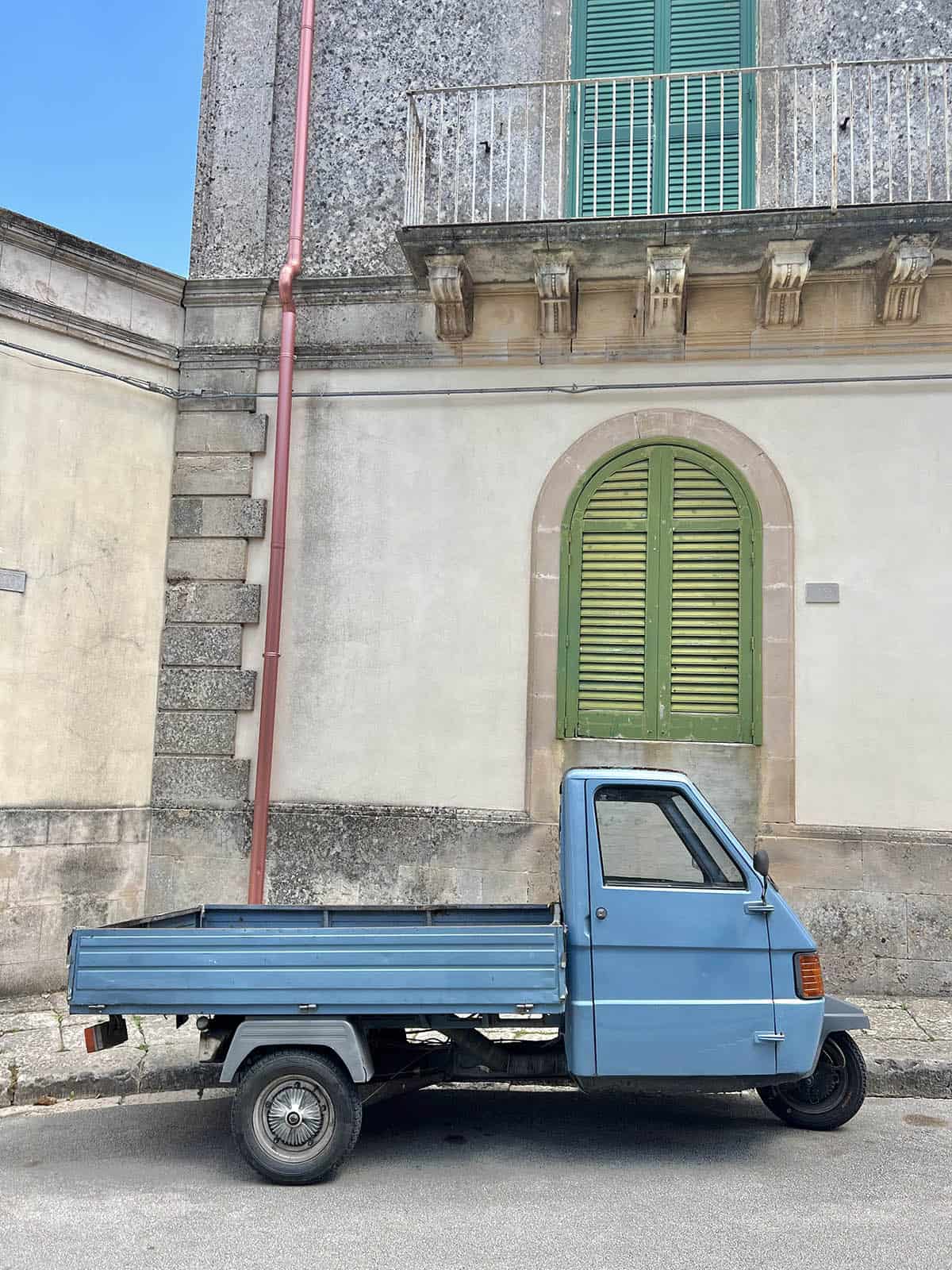 An image of a small blue car truck against the pastel streetscape of Ragusa Sicily