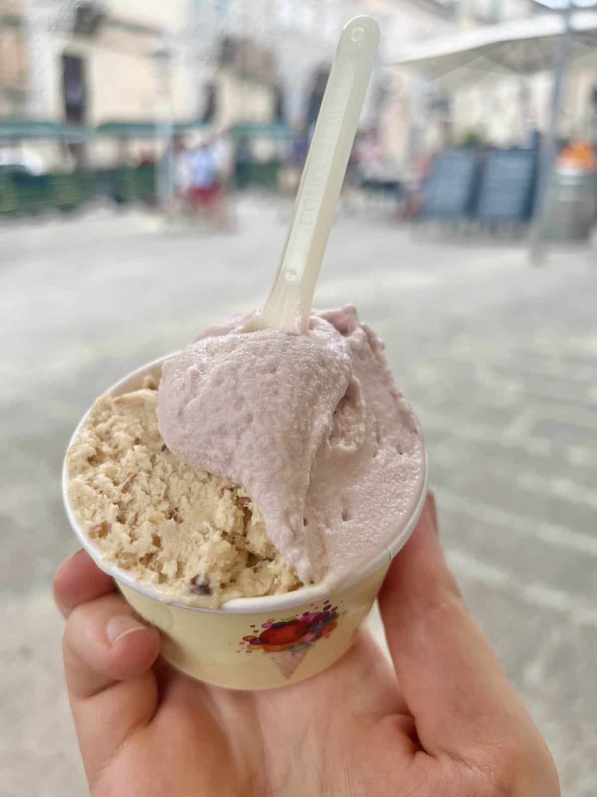 An image of a hand holding a cup of gelato from Gelati DiVini in Ragusa, Sicily. The streetscape of Ragusa Ibla is in the background