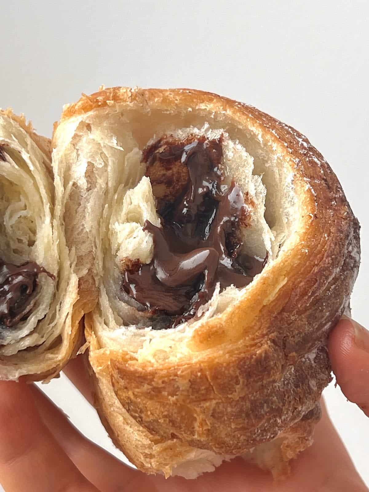 A close up image of a gluten free cornetto filled with Nutella that has been torn in half to reveal the chocolate insides. A hand holds both sides of the cornetto against a white wall.