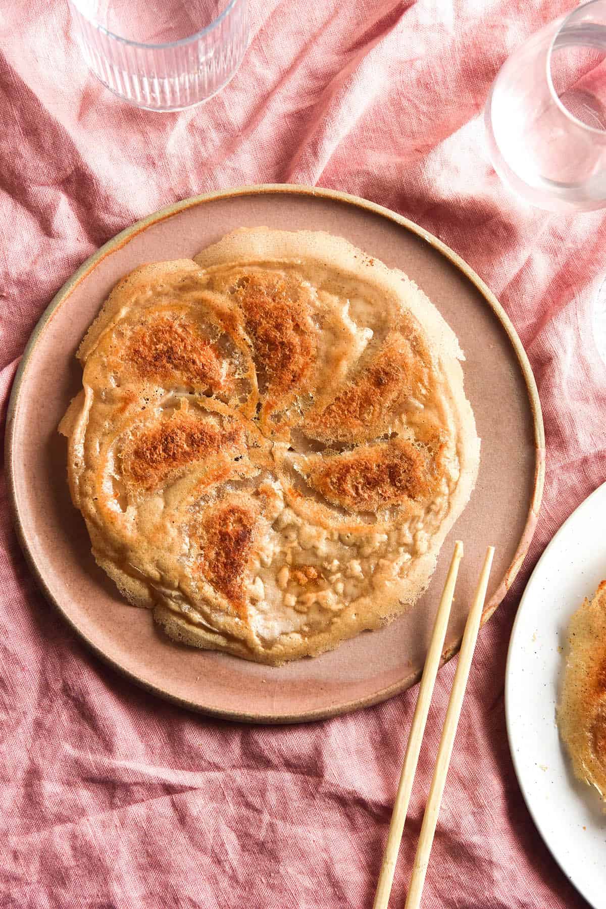 An image of gluten free skirt dumplings on a pale pink ceramic plate atop a pale pink linen tablecloth. A set of chopsticks sit to the bottom left of the plate. 