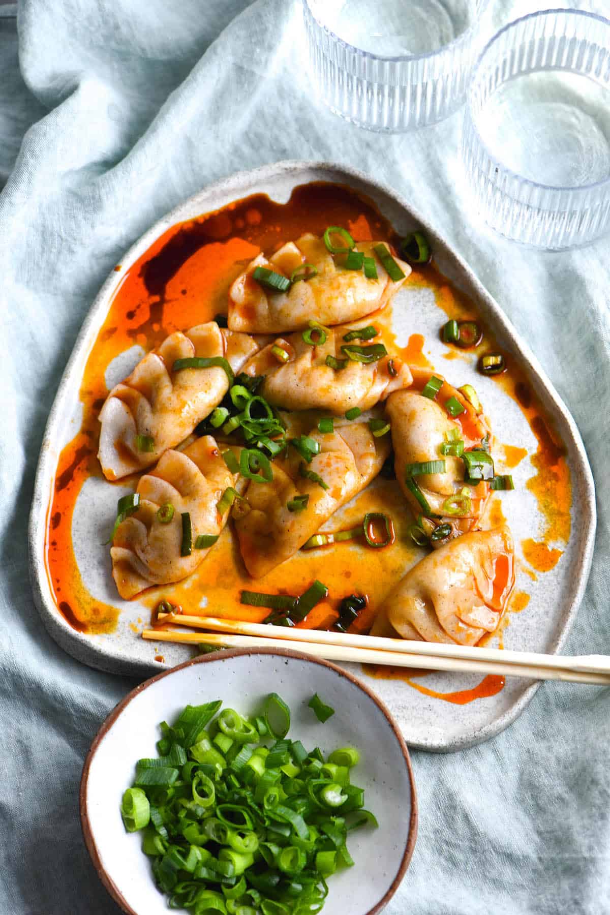 A plate of vegan, gluten free and FODMAP friendly dumplings sit atop a light green linen tablecloth. The dumplings are pleated, and drizzled with chilli oil, tamari and spring onion greens. Chopsticks rest on the edge of the plate and a small ceramic dish of spring onion greens sits to the bottom left of the dumplings