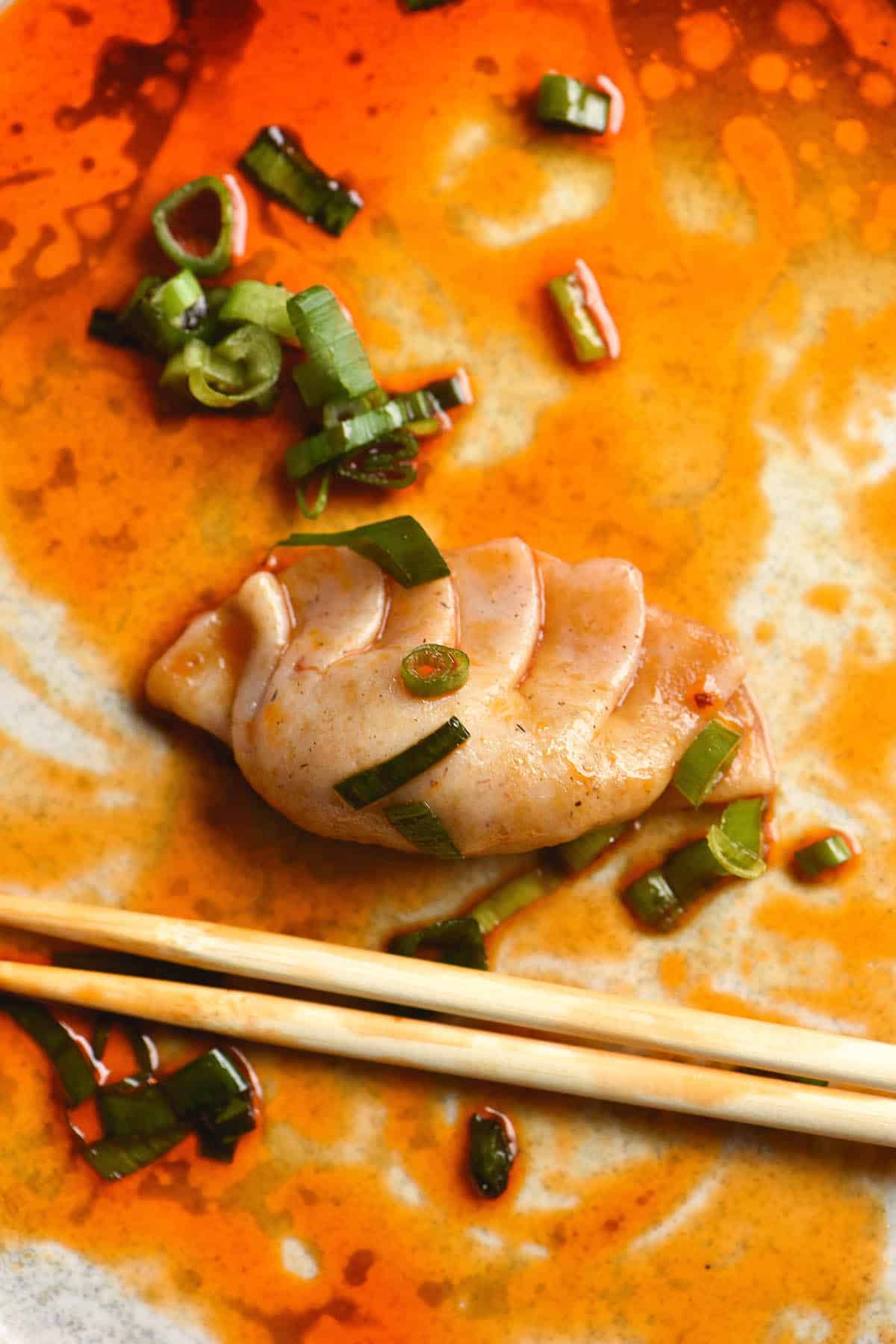 A close up image of a pleated gluten free dumpling smothered in chill oil on a white plate. Chopsticks sit underneath the dumpling.