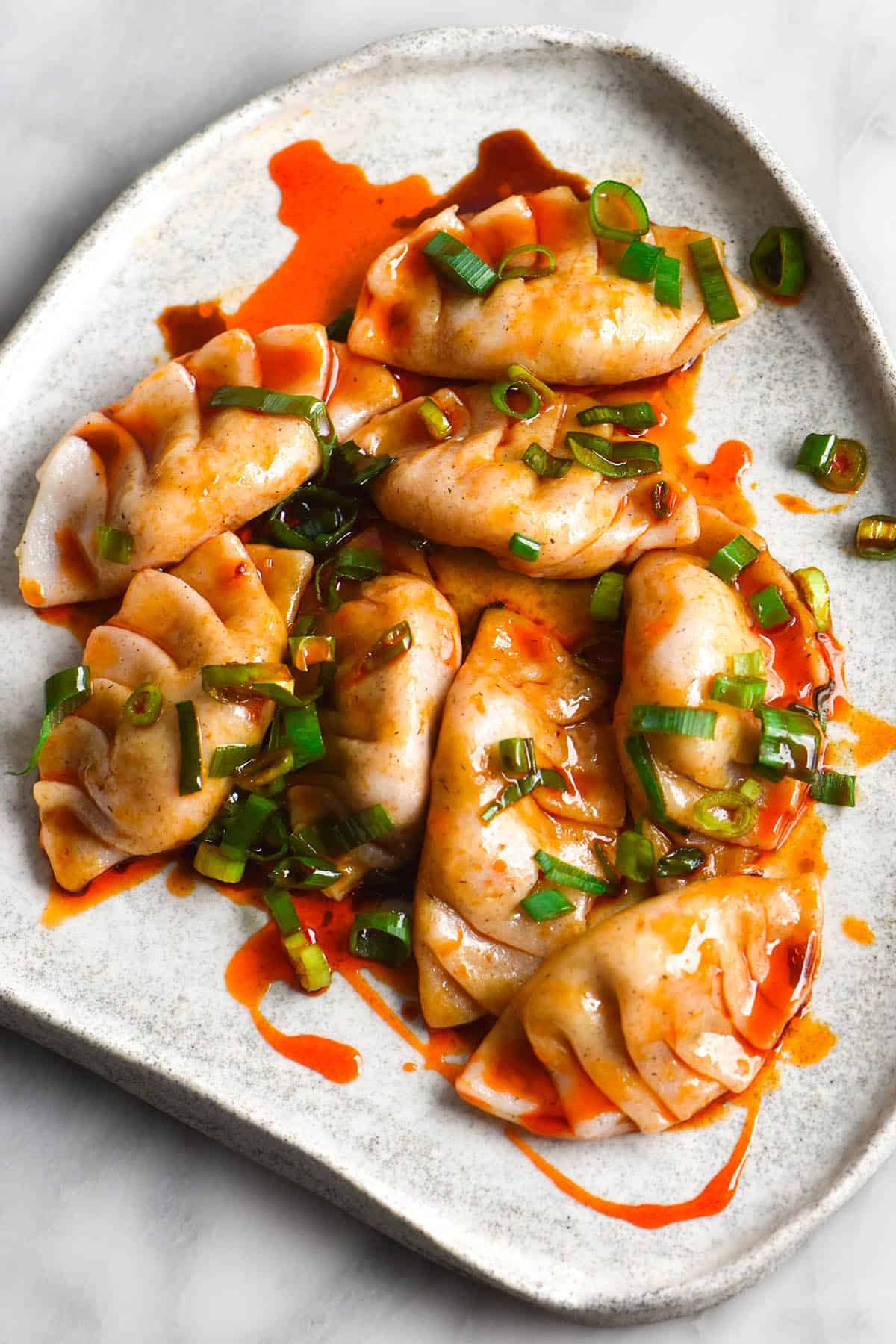 A close up image of pleated gluten free dumplings smothered in a deep red chilli sauce and finished with spring onion greens. The dumplings sit atop a white ceramic plate on a white marble table. 