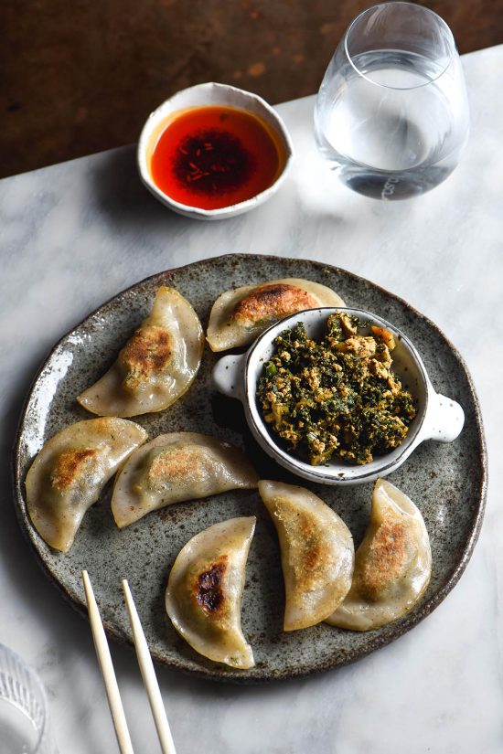 An aerial side on image of a plate of pan fried gluten free dumplings with a tofu and bok choi dumpling filling. The plate sits atop a white marble table and a small bowl of chilli oil sits in the top right corner.