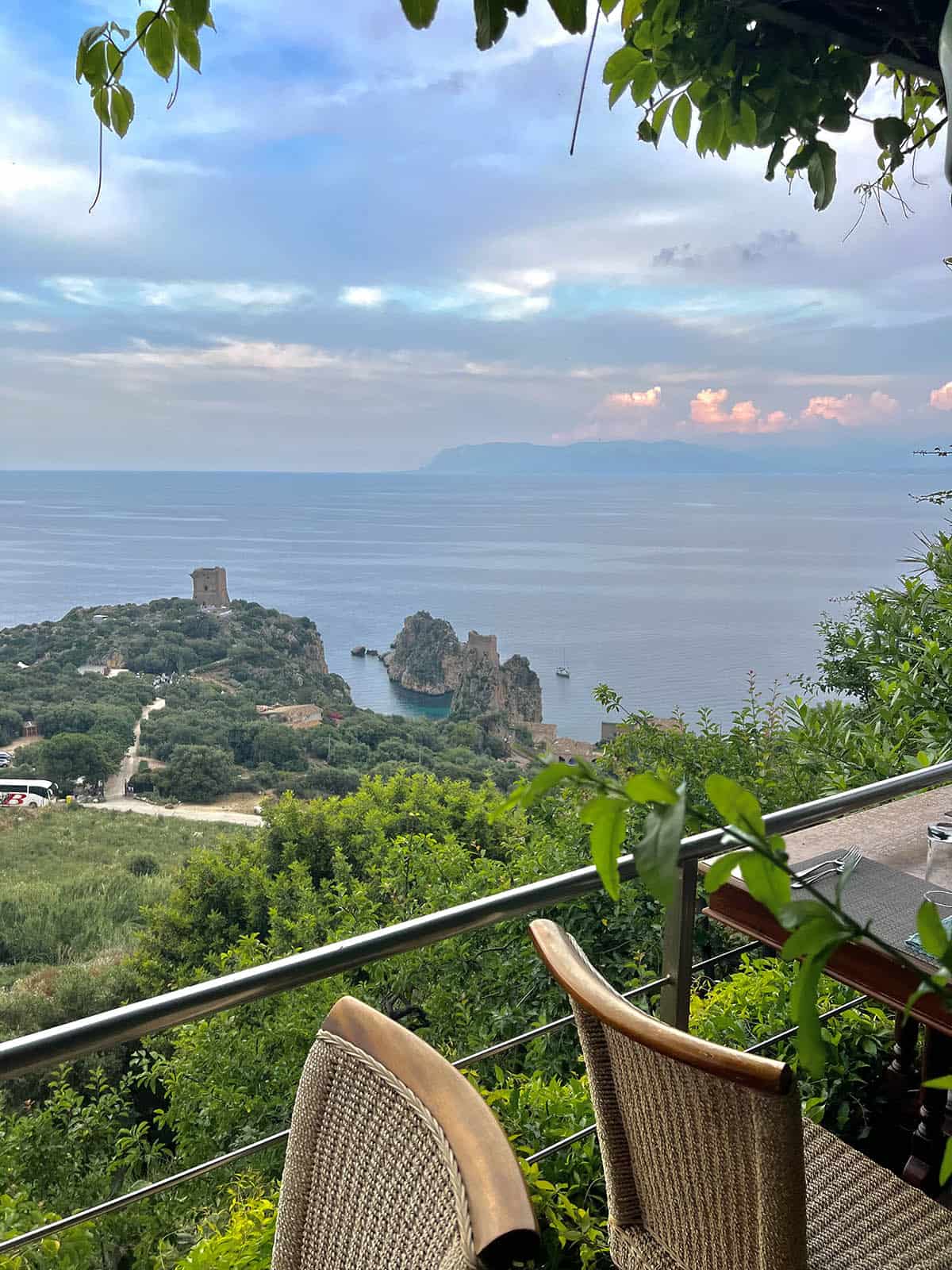 An image of the view of Scopello and Tonnara Di Scopello from Hotel Bennistra Ristorante. The blue ocean and lush green landscape sit in the background while the restaurant tables sit perched up the hill in the foreground.