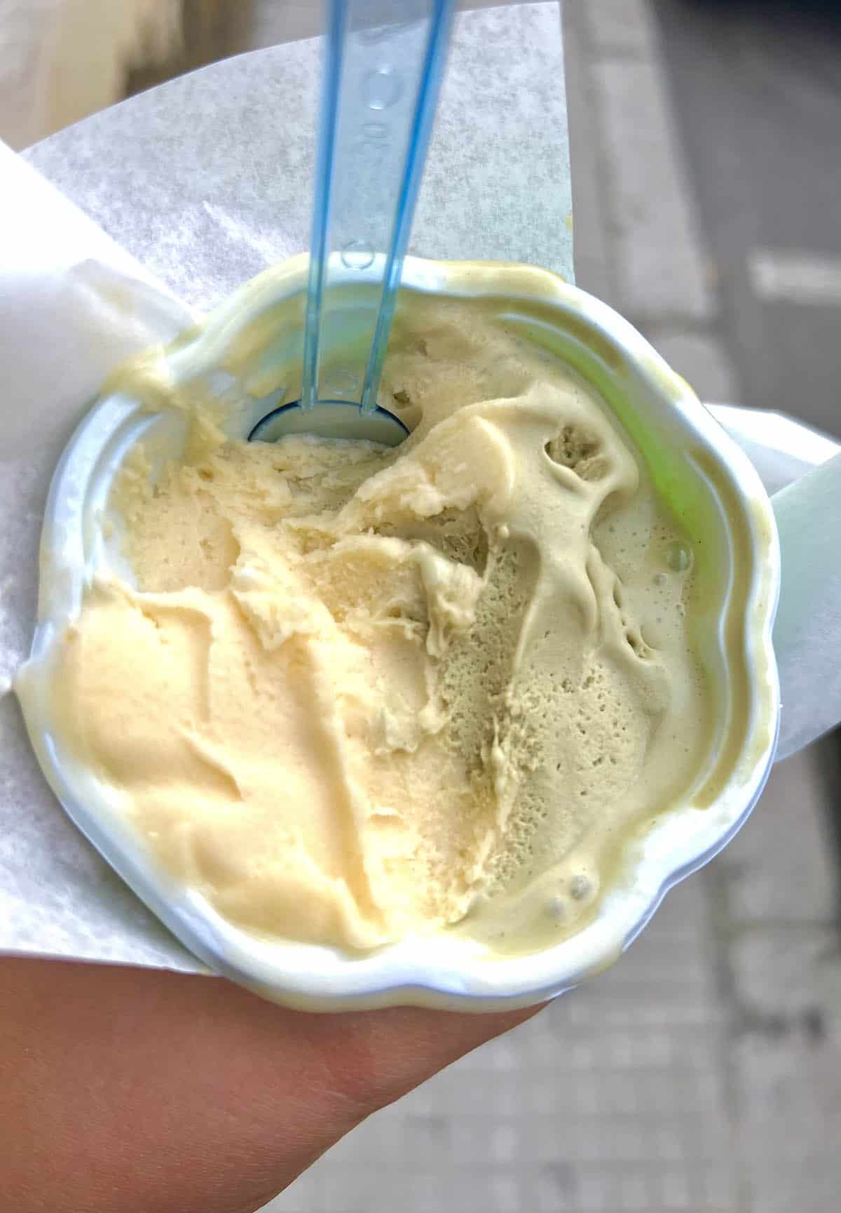 An image of a hand holding a peach and pistachio flavoured gelato in a cup at the roadside in Sicily. 