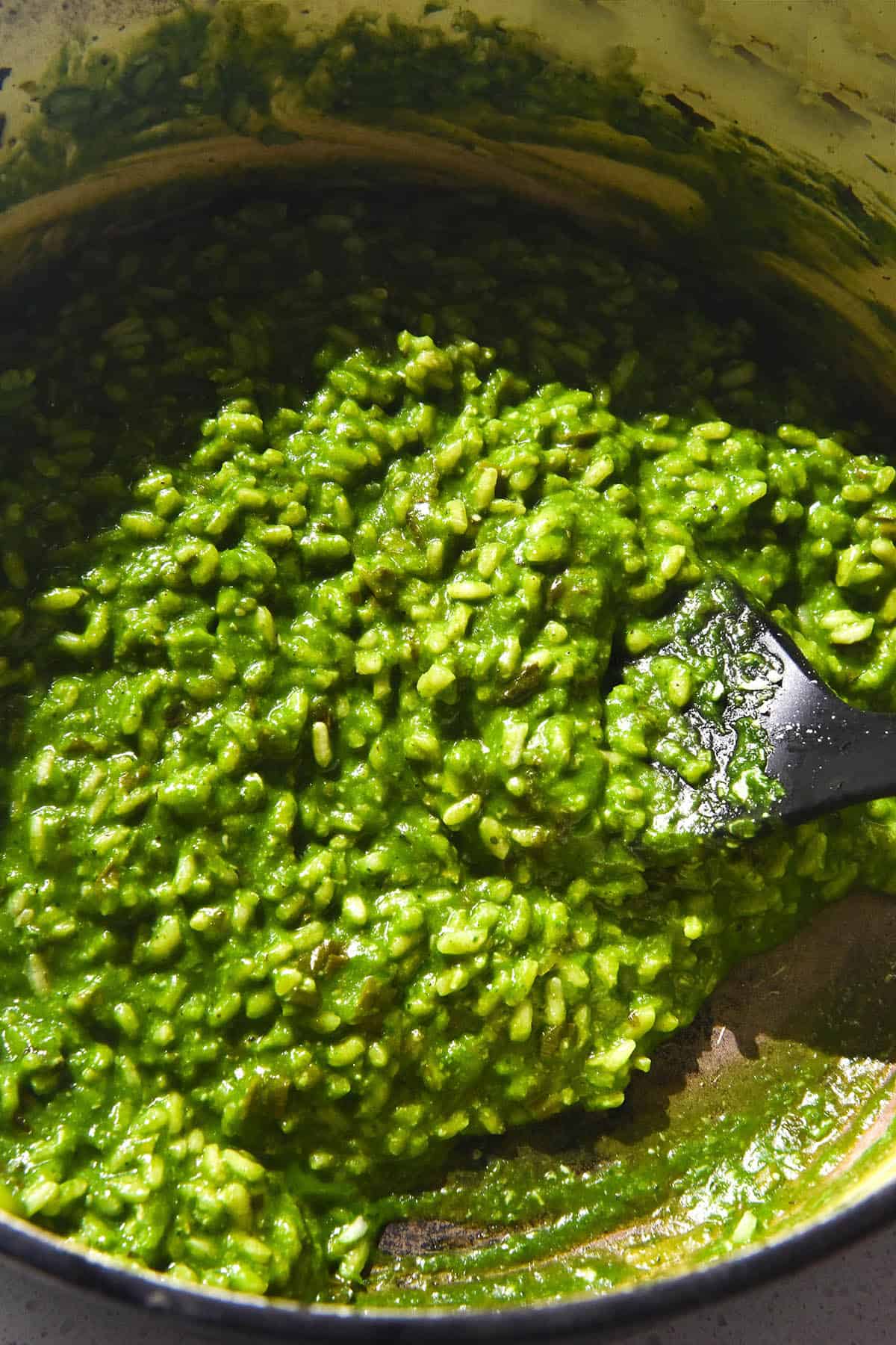 A brightly lit image of a dutch oven filled with bright green risotto verde. A spatula sits in the risotto on the right hand side of the image. 
