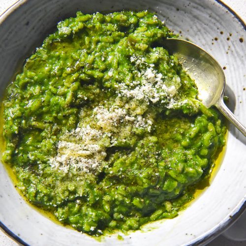 A brightly lit aerial image of a white ceramic bowl filled with risotto verde. The risotto is topped with finely grated parmesan, freshly cracked pepper and some olive oil. The bowl sits atop a white stone kitchen counter.
