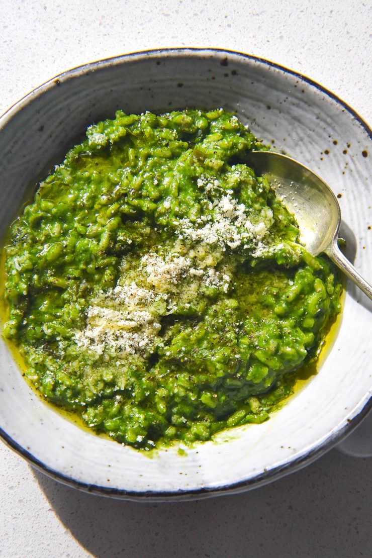 A brightly lit aerial image of a white ceramic bowl filled with risotto verde. The risotto is topped with finely grated parmesan, freshly cracked pepper and some olive oil. The bowl sits atop a white stone kitchen counter.