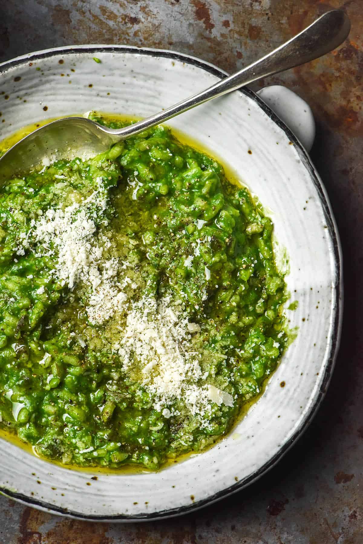 A close up image of a white ceramic bowl filled with low FODMAP risotto verde topped with grated parmesan and olive oil. A spoon sticks into the risotto from the top right of the image. The bowl sits atop a dark grey rusty coloured backdrop. 