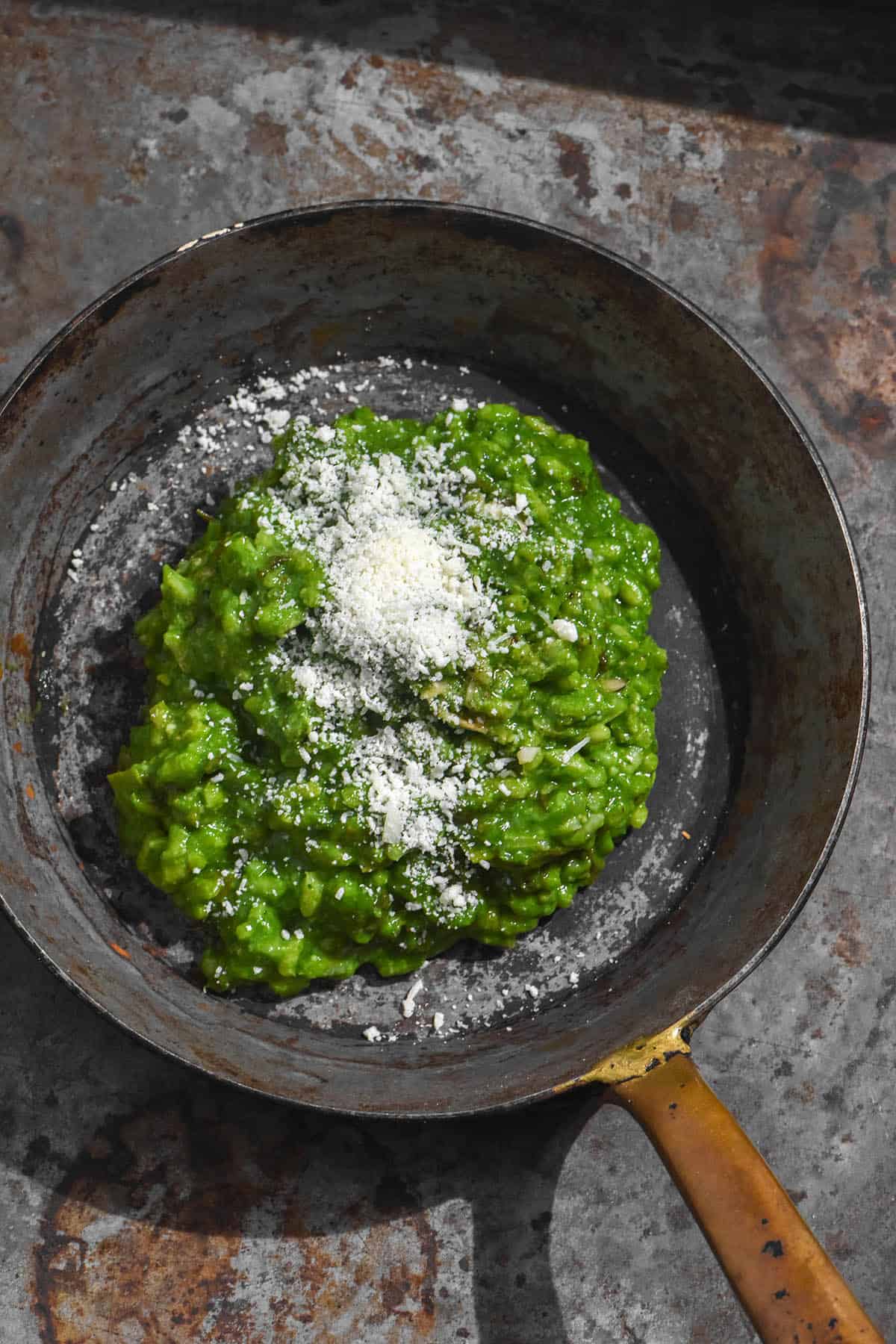 A brightly lit aerial image of low FODMAP risotto verde in a small vintage grey skillet atop a grey rusted steel backdrop. The risotto is bright green and topped with freshly grated parmesan.
