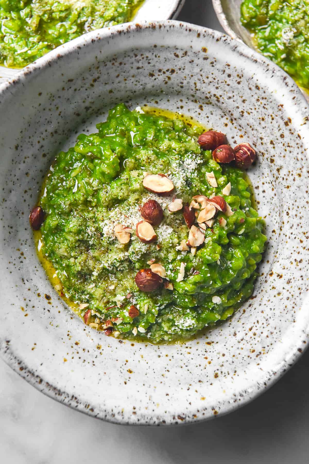 An aerial close up image of a white speckled ceramic bowl filled with low FODMAP risotto verde. The risotto is bright green and topped with parmesan, olive oil and chopped hazelnuts. 