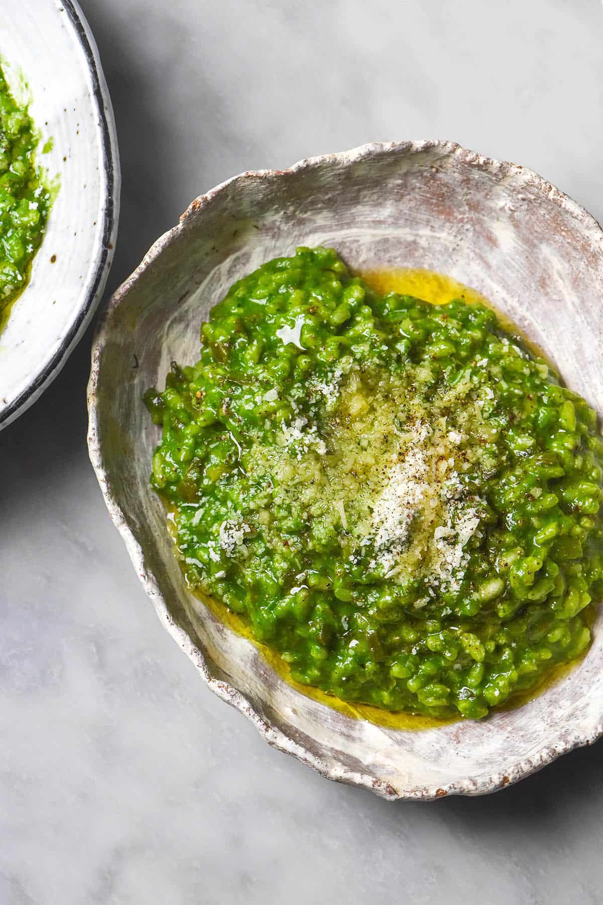 An aerial close up image of a white rustic ceramic bowl filled with risotto verde. The risotto verde is bright green and is topped with parmesan and olive oil. The bowl sits atop a white marble table and a second bowl sits off to the top left of the image. 