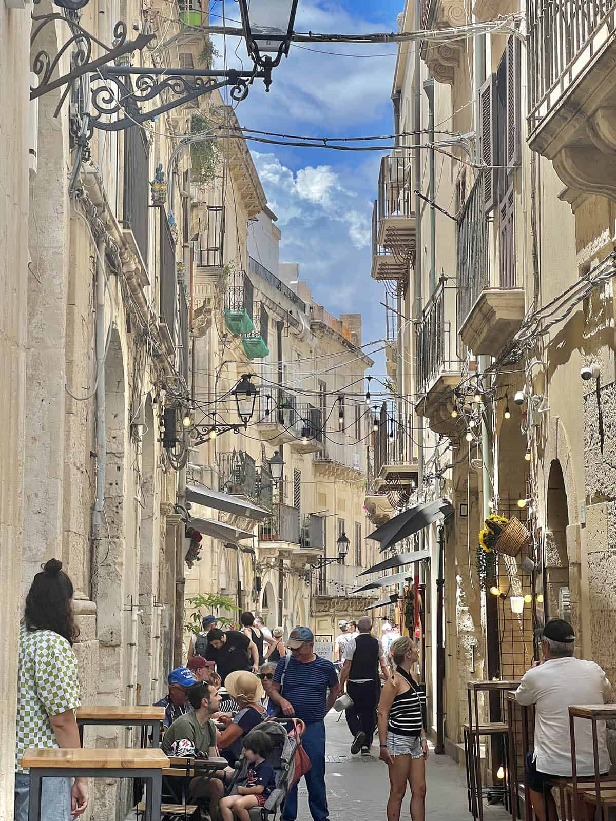 A streetscape in Ortigia, Sicily filled with people eating in restaurants and blue skies. Fairy lights and old lamps adorn the shops.
