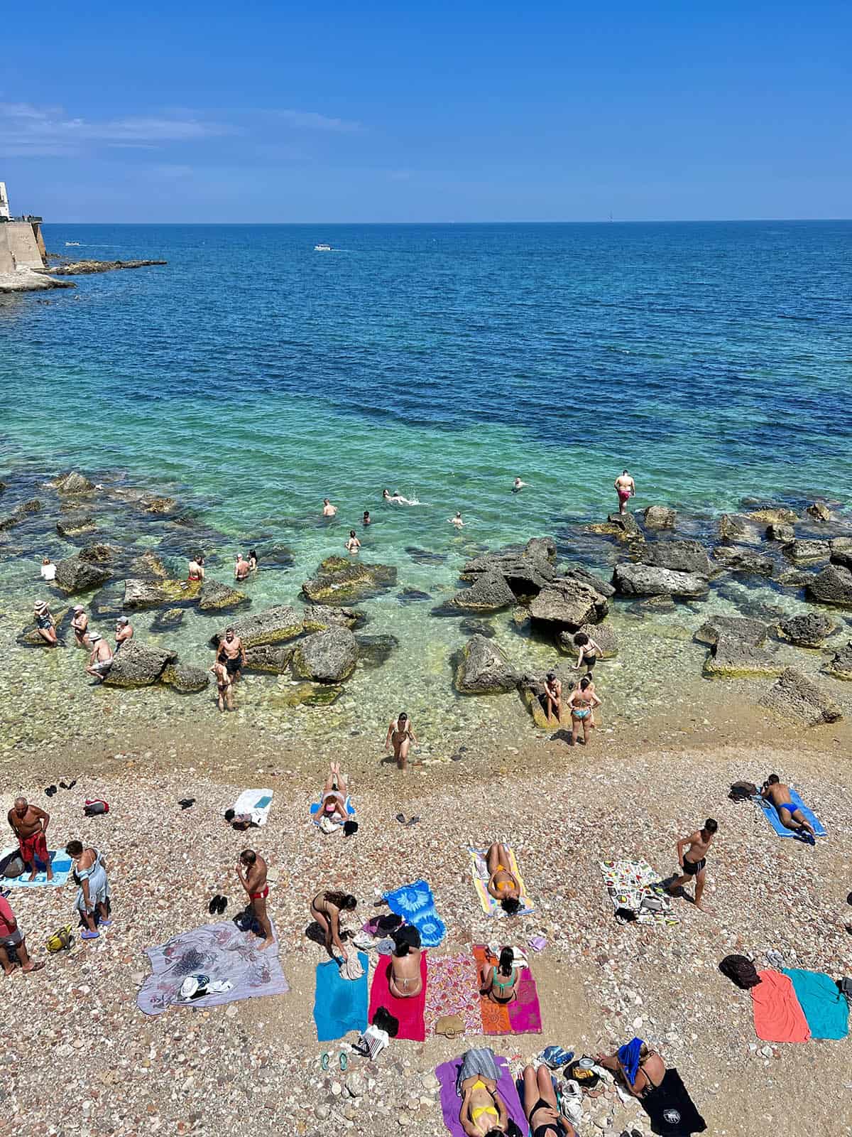 An image of the aquamarine blue waters and summer bathers at Cala Rossa in Ortigia