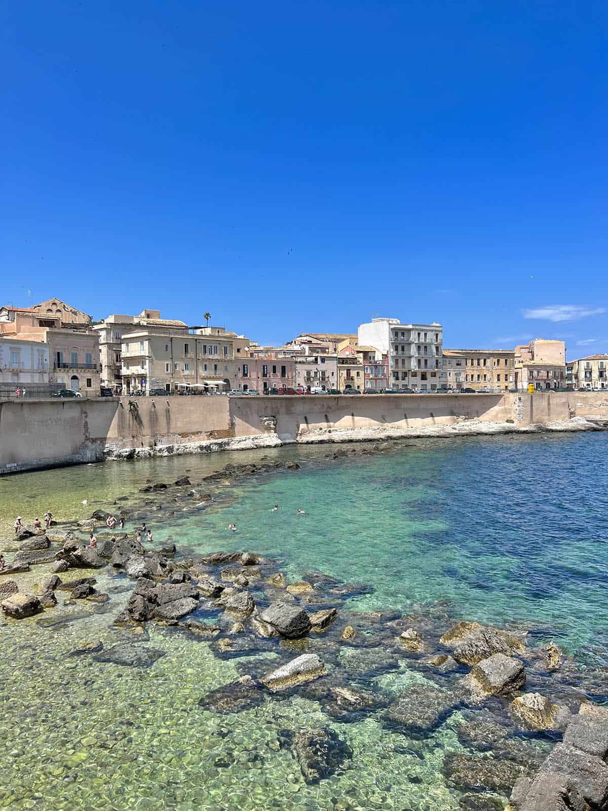 An image of the coastline of Ortigia with aquamarine waters, tan coloured buildings and a bright blue sky. 