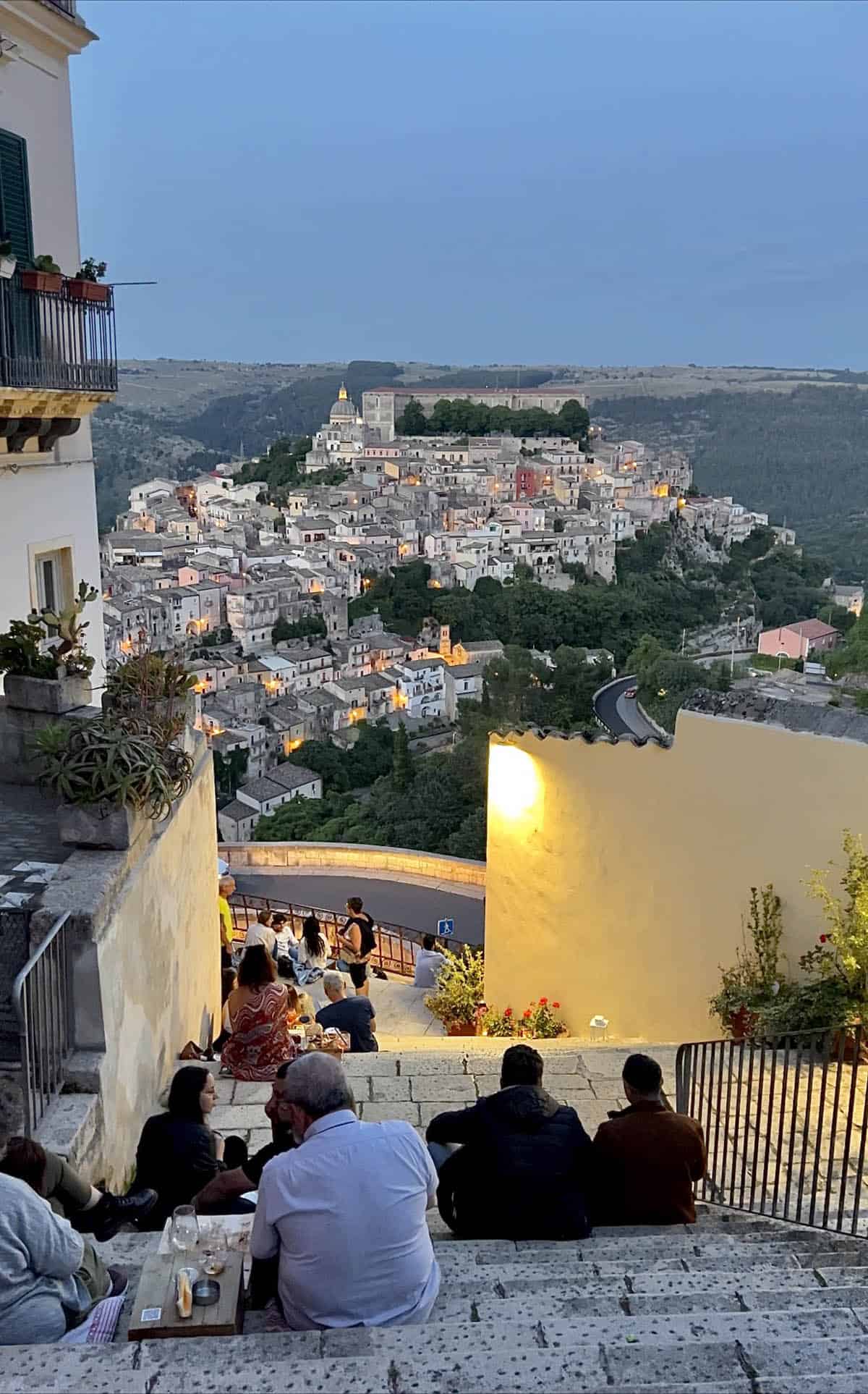 An image of people sitting on the stairs at Al Gradino 284 in Ragusa Superior overlooking the sun setting over Ragusa Ibla