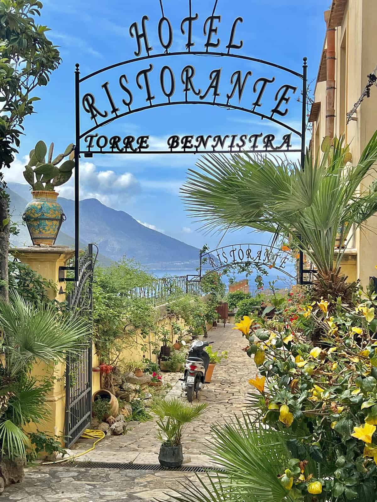 An image of the hotel Torre Bennistra with the mountains and ocean of Scopello in the background. Lush greenery in the foreground frames the hotel sign and the bright blue sky