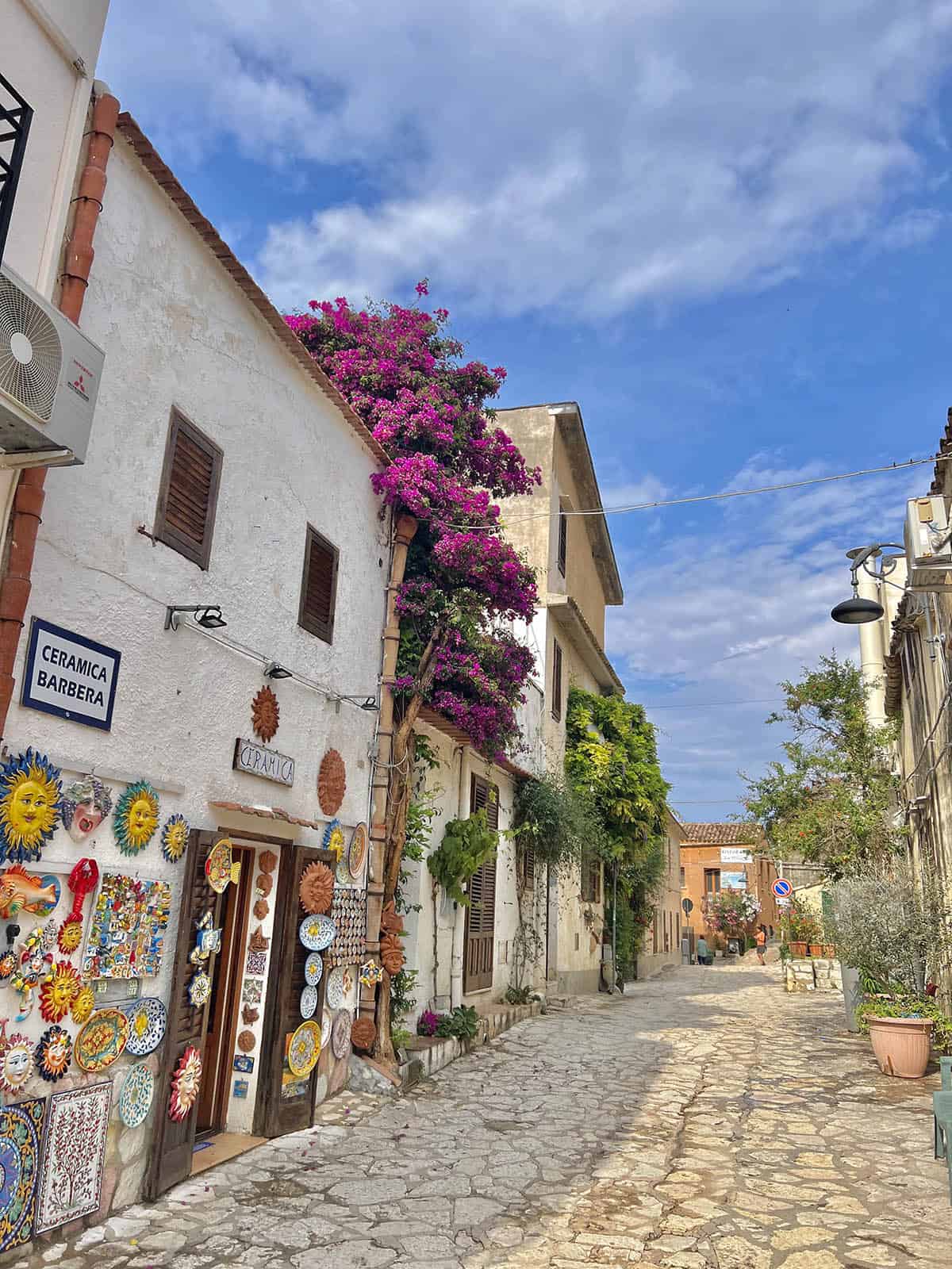 An image of the cobbled streets of Scopello, Sicily. In the foreground is a ceramic shop with a large bougainvillea tree on the balcony. The sky is blue and sunny
