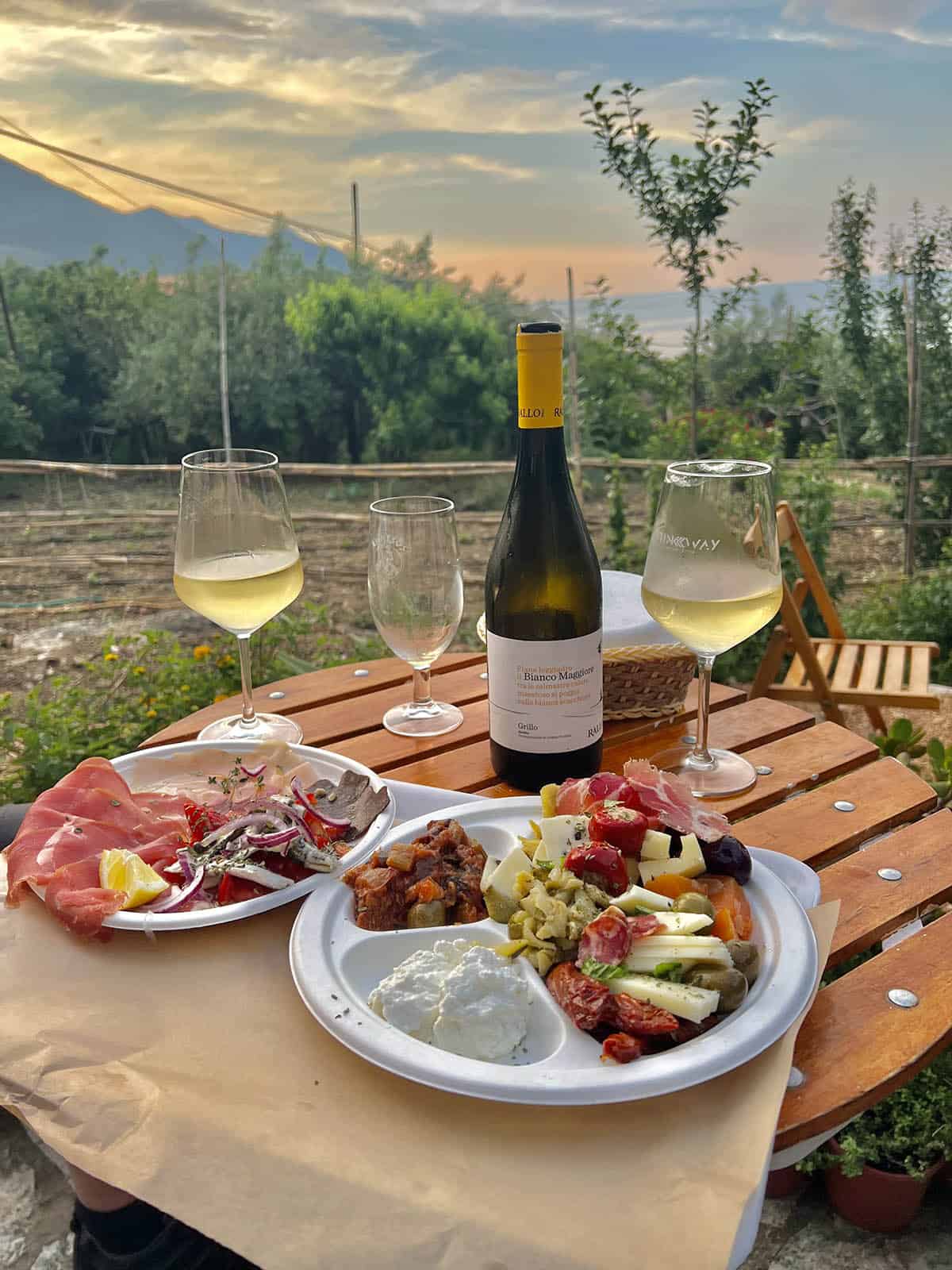 An image of Sicilian antipasto plates, white wine and wine glasses on a table in a garden in Scopello, Sicily. The image has been taken at sunset and the mountains and ocean are visible in the background. 