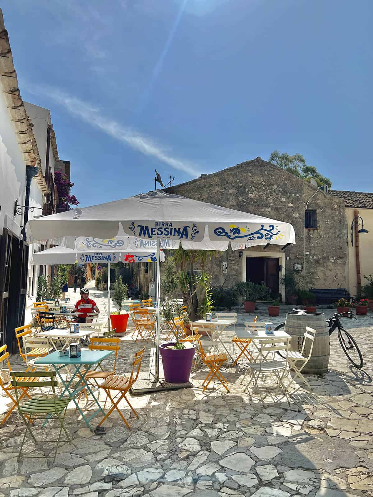 An image of the square in Scopello, Sicily. Coffee tables, chairs and umbrellas sit in a cobbled courtyard on a sunny summer morning. 