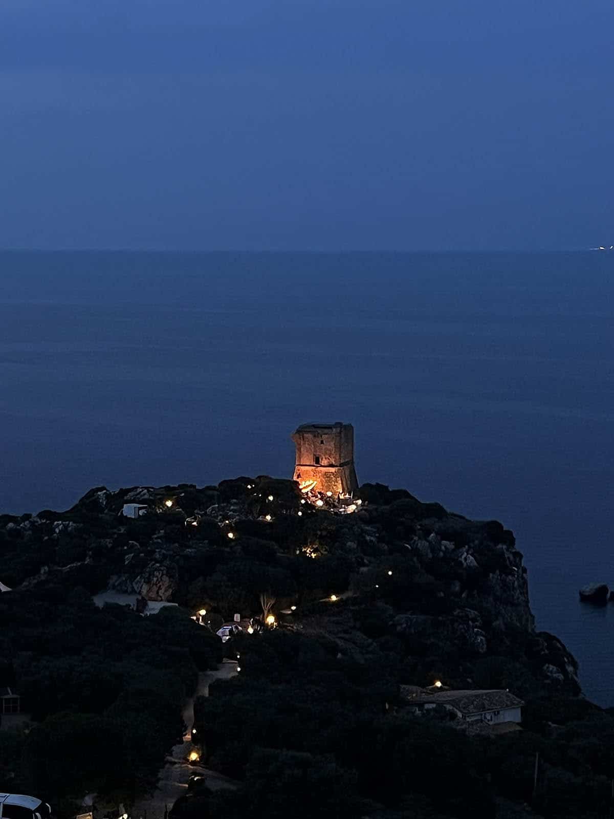A nighttime image of a wedding at Torre Di Scopello in Scopello Sicily. Lights illuminate the tower and fairy lights light up the wedding below. 