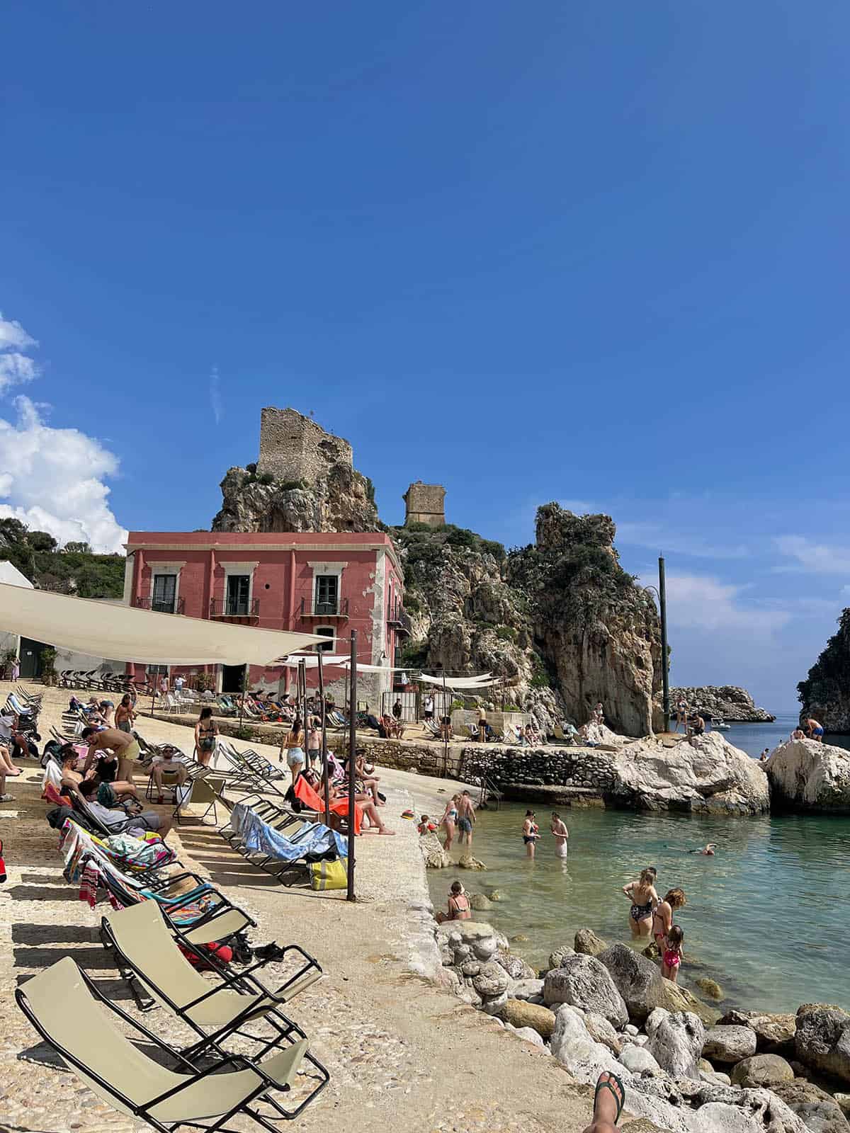 An image of Tonnara Di Scopello on a warm summer's day. Shade sails and seating are up and people swim in the aquamarine water. 