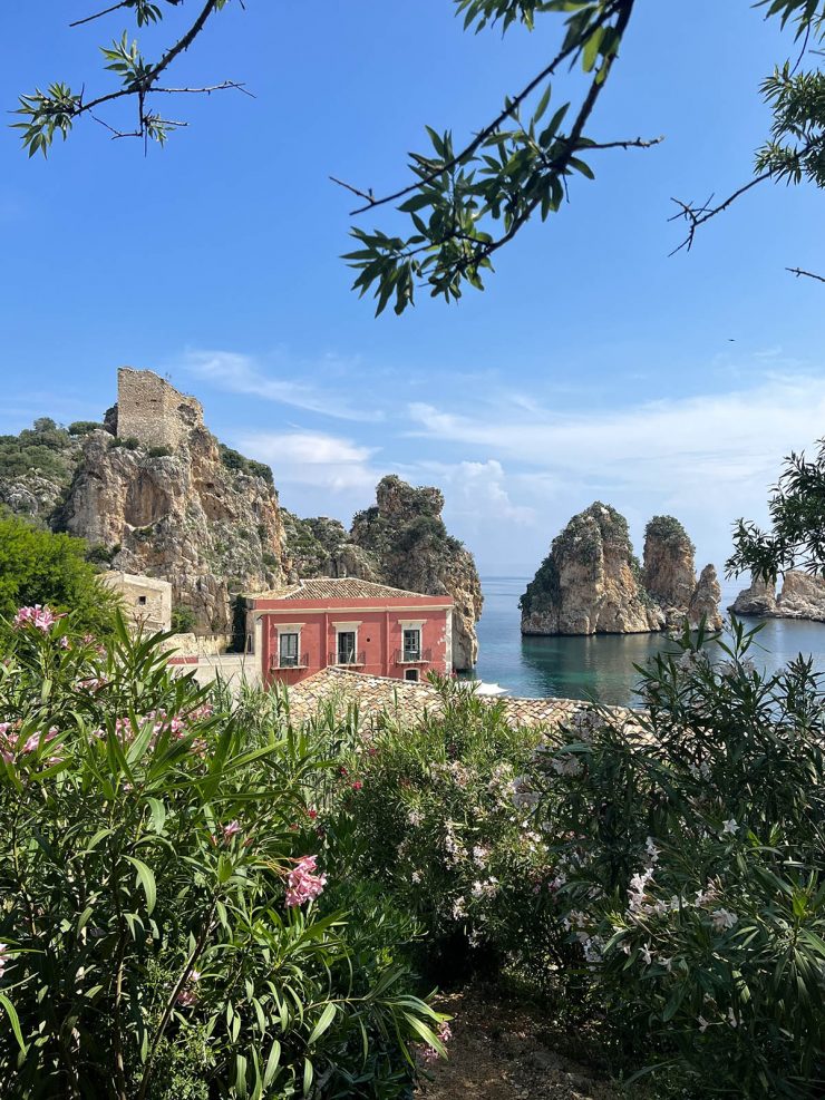 An image of the Tonnara and Torre Di Scopello with the ocean in the background and lush green plants in the foreground