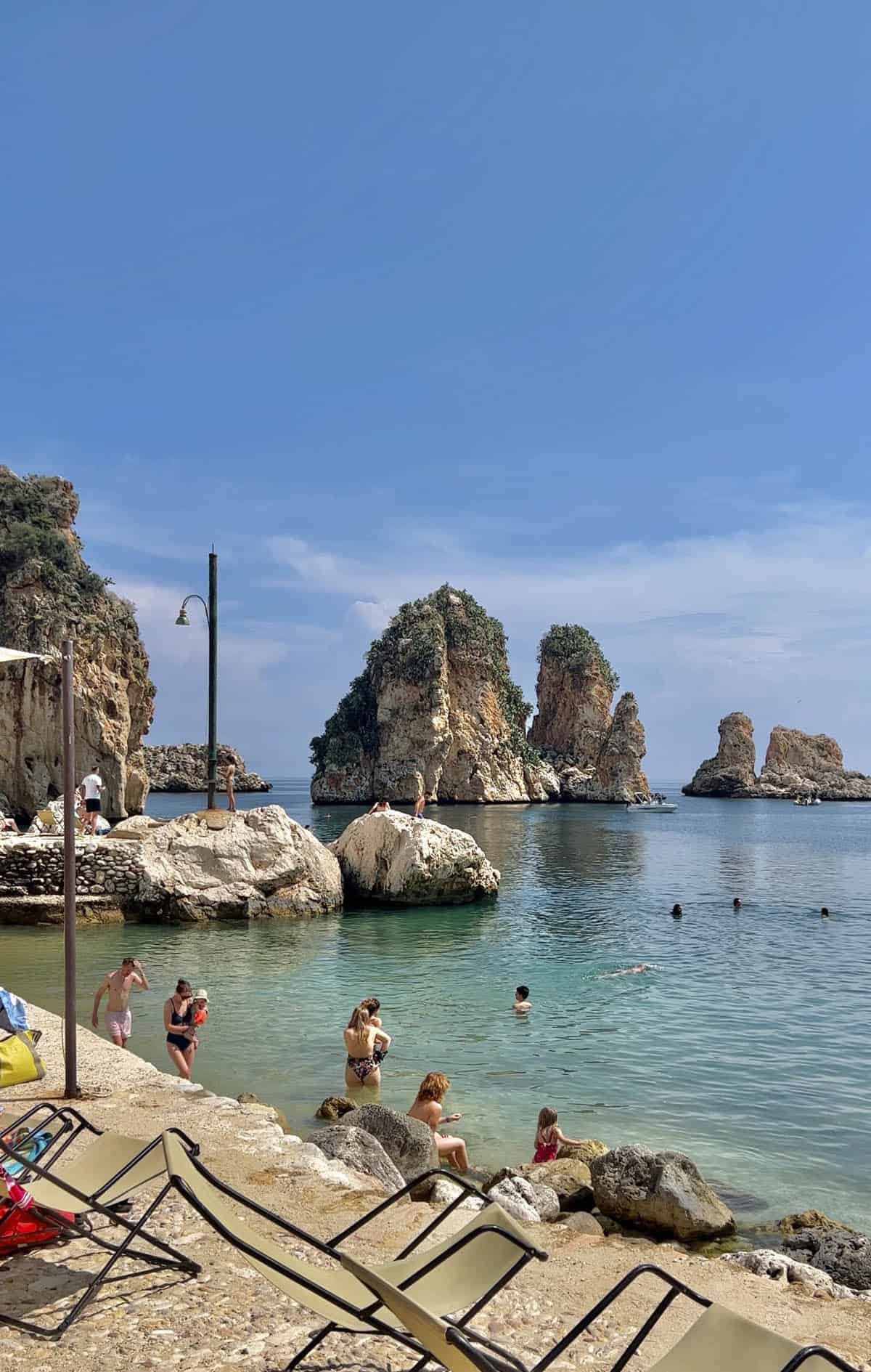 An image of the aquamarine waters, jagged rocks, sunbeds and bathers of Tonnara di Scopello on a bright summer day