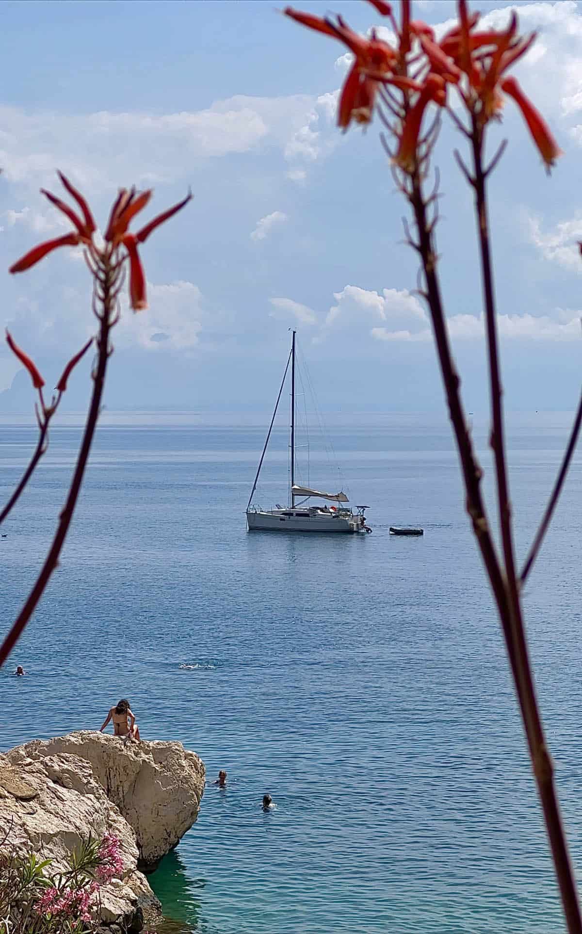A view of a yacht in the waters surrounding Scopello framed with long stemmed flowers. A rock juts out from the bottom left hand corner and a woman sits on the rock. People swim in the aquamarine waters surrounding the rock. 