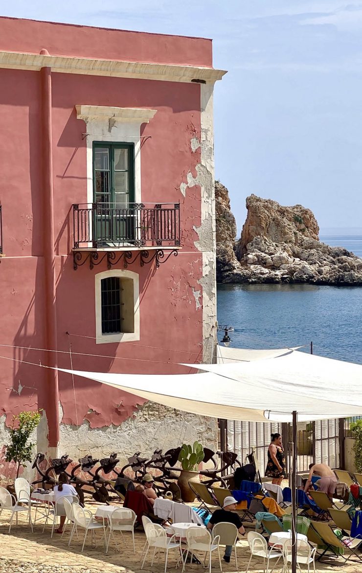 An image of the Tonnara Di Scopello in summer. Shade sails protect the throng of bathers on sun loungers next to the pink building, and the ocean is in the background.