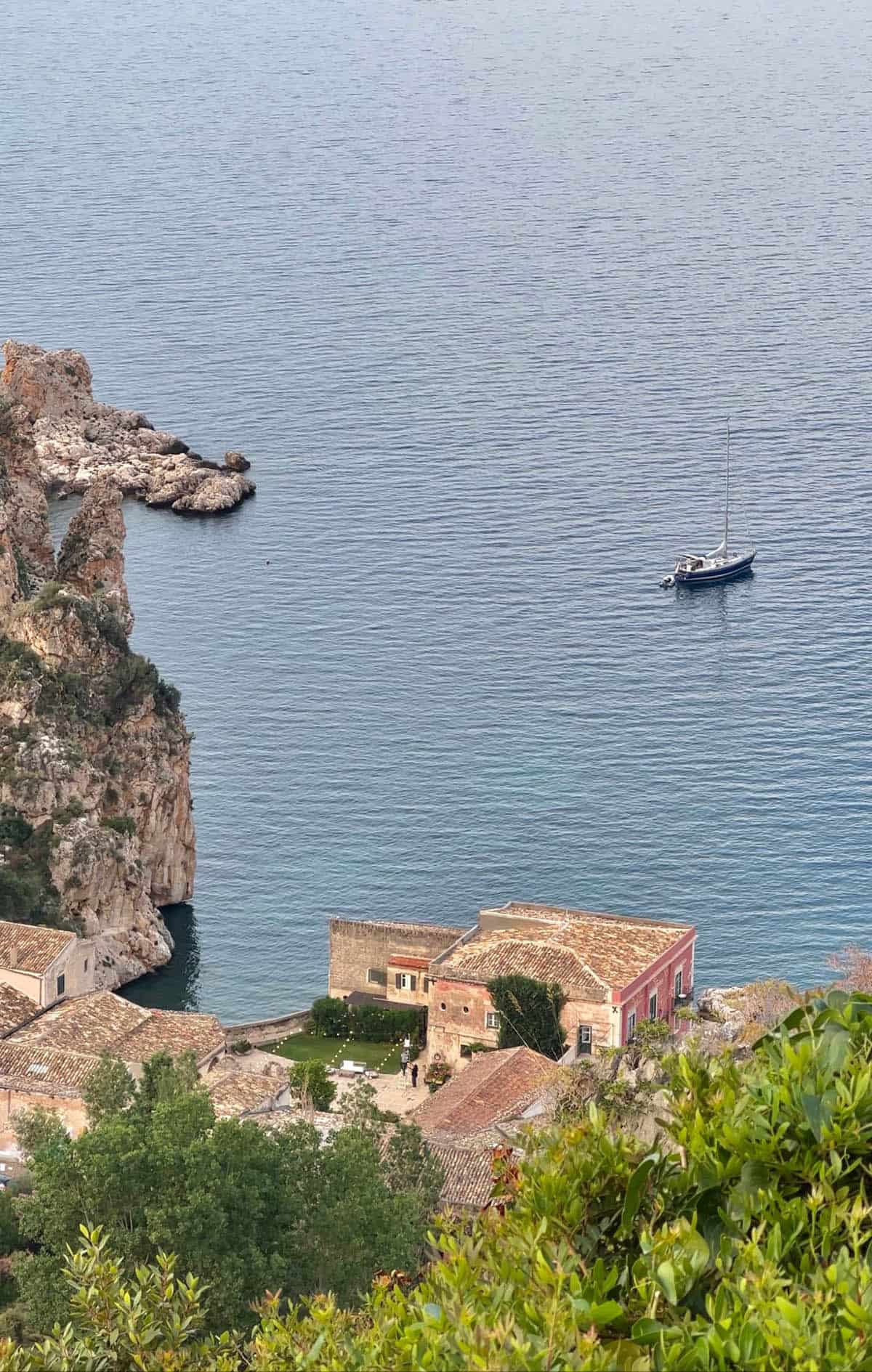 The view of La Tonnara Di Scopello from La Terrazza restaurant in Scopello. The Tonnara sits on the edge of the waters and a yacht sits just offshore. Fairy lights are lit up in the courtyard of the Tonnara. 