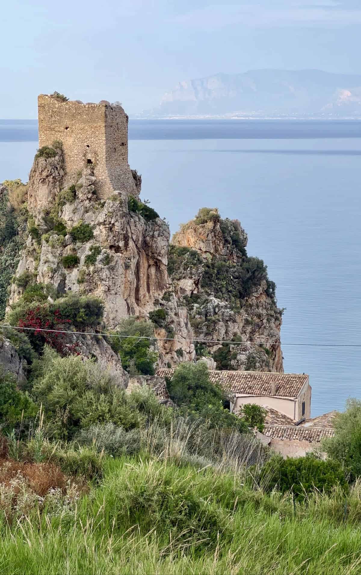 An image of the Torre Della Scopello and Tonnara Di Scopello with the sea as the backdrop. A mountainous cliff of Sicily is in the background. 