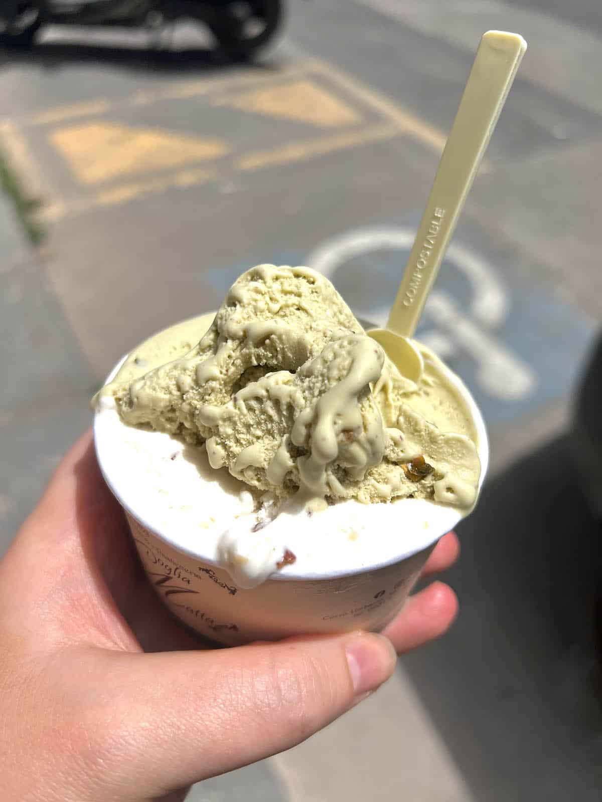 An image of a hand holding out a cup of gelato with stracciatella hazelnut and pistachio flavours by the roadside in Sicily.