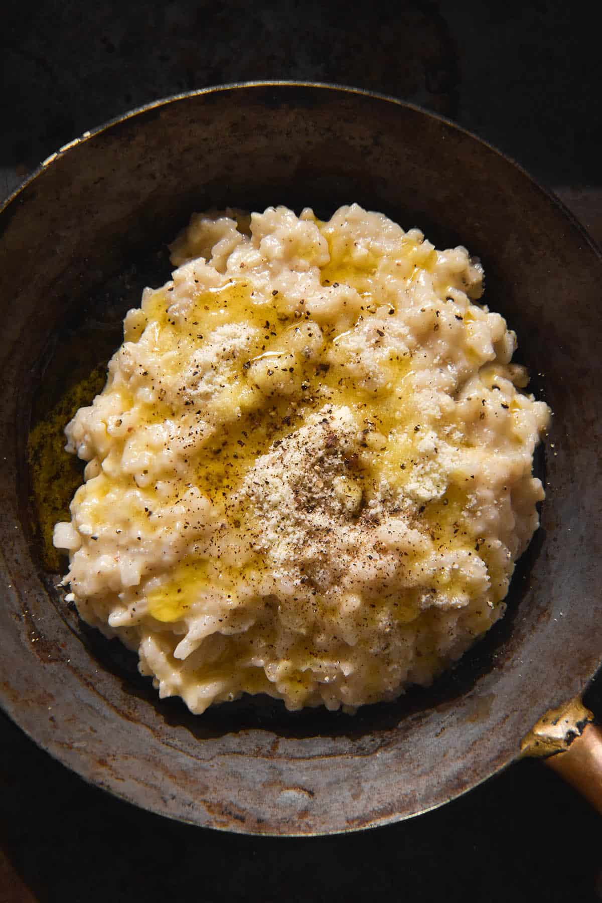A dark and moody image of a mini steel skillet filled with cacio e pepe risotto. The risotto is topped with extra parmesan and pepper and a drizzle of garlic infused ghee. 