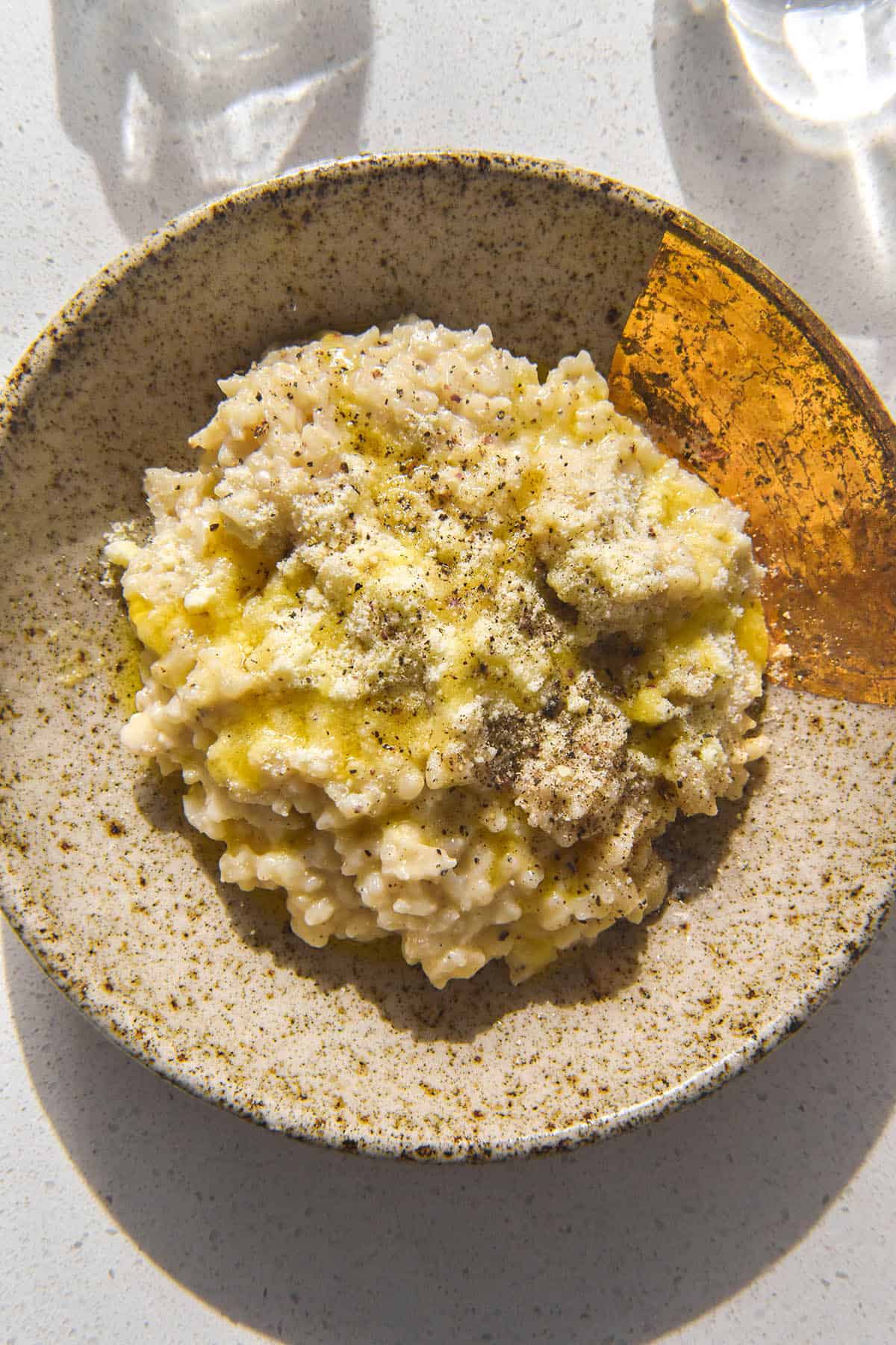 A brightly lit aerial image of cacio e pepe risotto in a speckled beige ceramic plate with a gold dipped corner. The risotto is topped with extra parmesan, pepper and a drizzle of garlic infused ghee. 