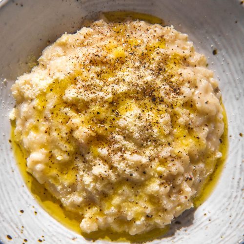 A close up aerial image of a white ceramic bowl of cacio e pepe risotto topped with extra parmesan, pepper and garlic infused ghee.