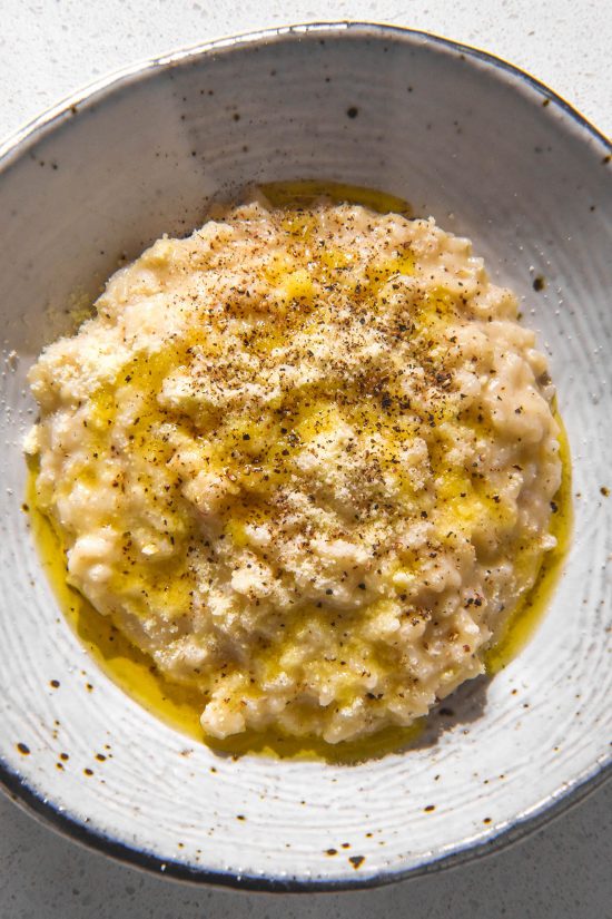 A close up aerial image of a white ceramic bowl of cacio e pepe risotto topped with extra parmesan, pepper and garlic infused ghee.