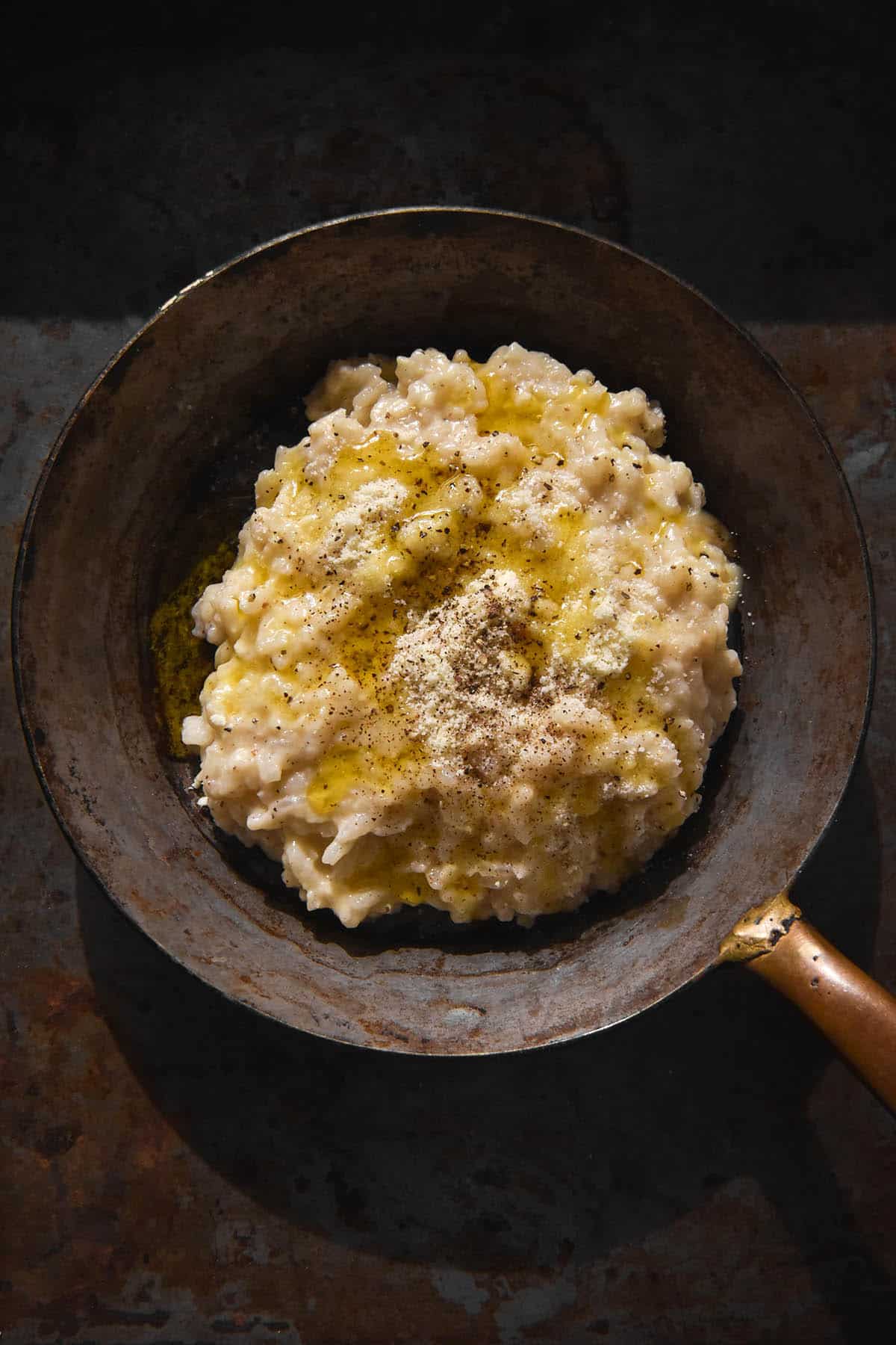 An aerial image of a rustic mini skillet filled with cacio e pepe risotto topped with extra cheese, pepper and a drizzle of garlic infused ghee. The skillet sits on a rustic grey backdrop. 