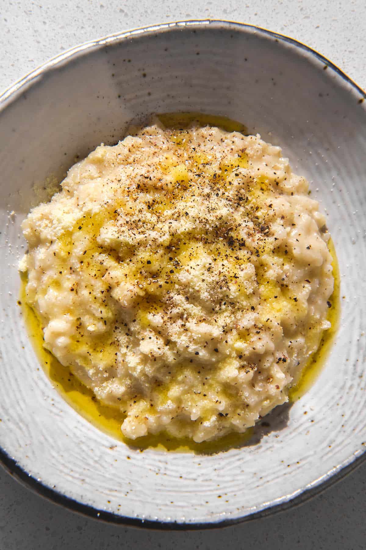 An aerial image of a white textured ceramic bowl filled with cacio e pepe risotto. The risotto is finished with extra cheese, pepper and a drizzle of garlic infused ghee. 
