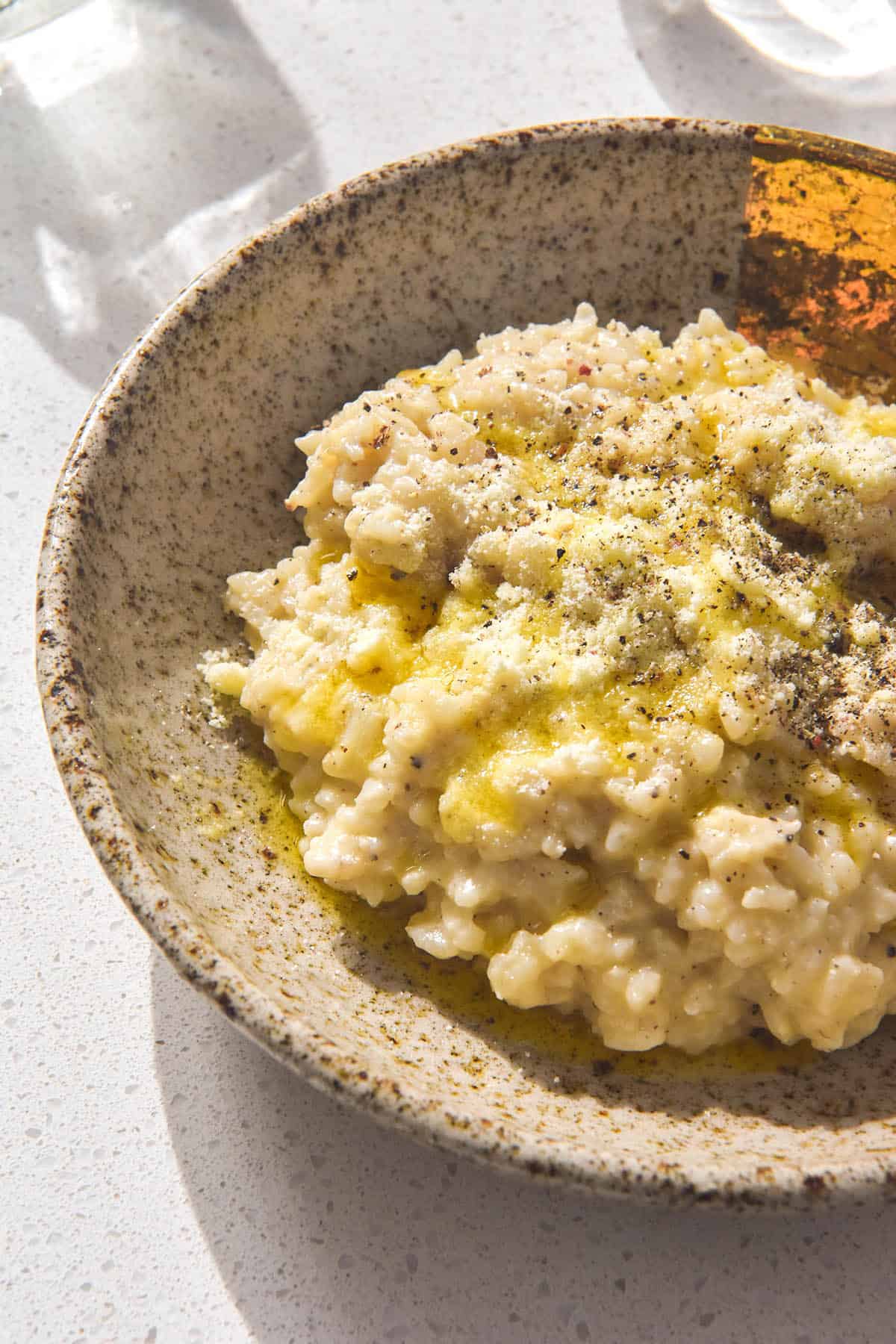 A brightly lit side on image of a beige speckled ceramic bowl filled with cacio e pepe risotto. The risotto is topped with extra cheese, pepper and a drizzle of garlic infused ghee. It sits on a brightly lit white stone bench top and two glasses of water sit in the background. 